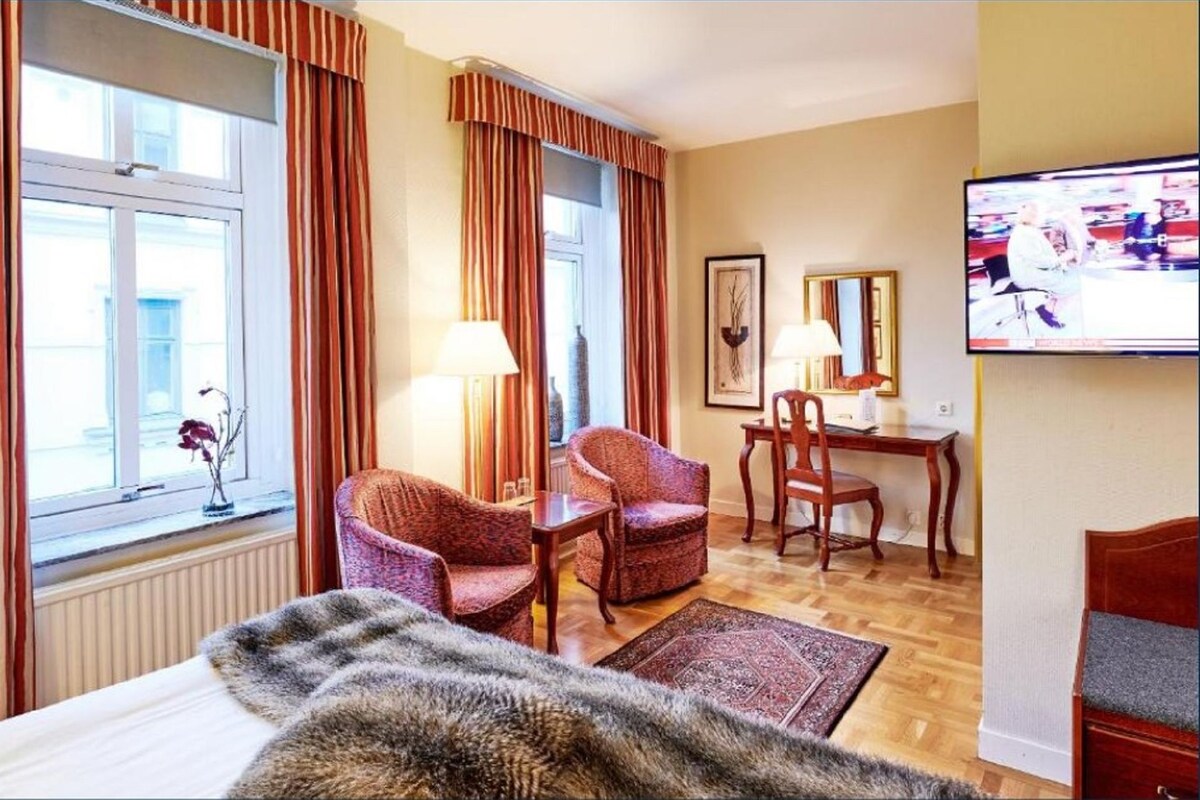 A hotel room with a bed, two chairs, a desk, a television, and a window with curtains.