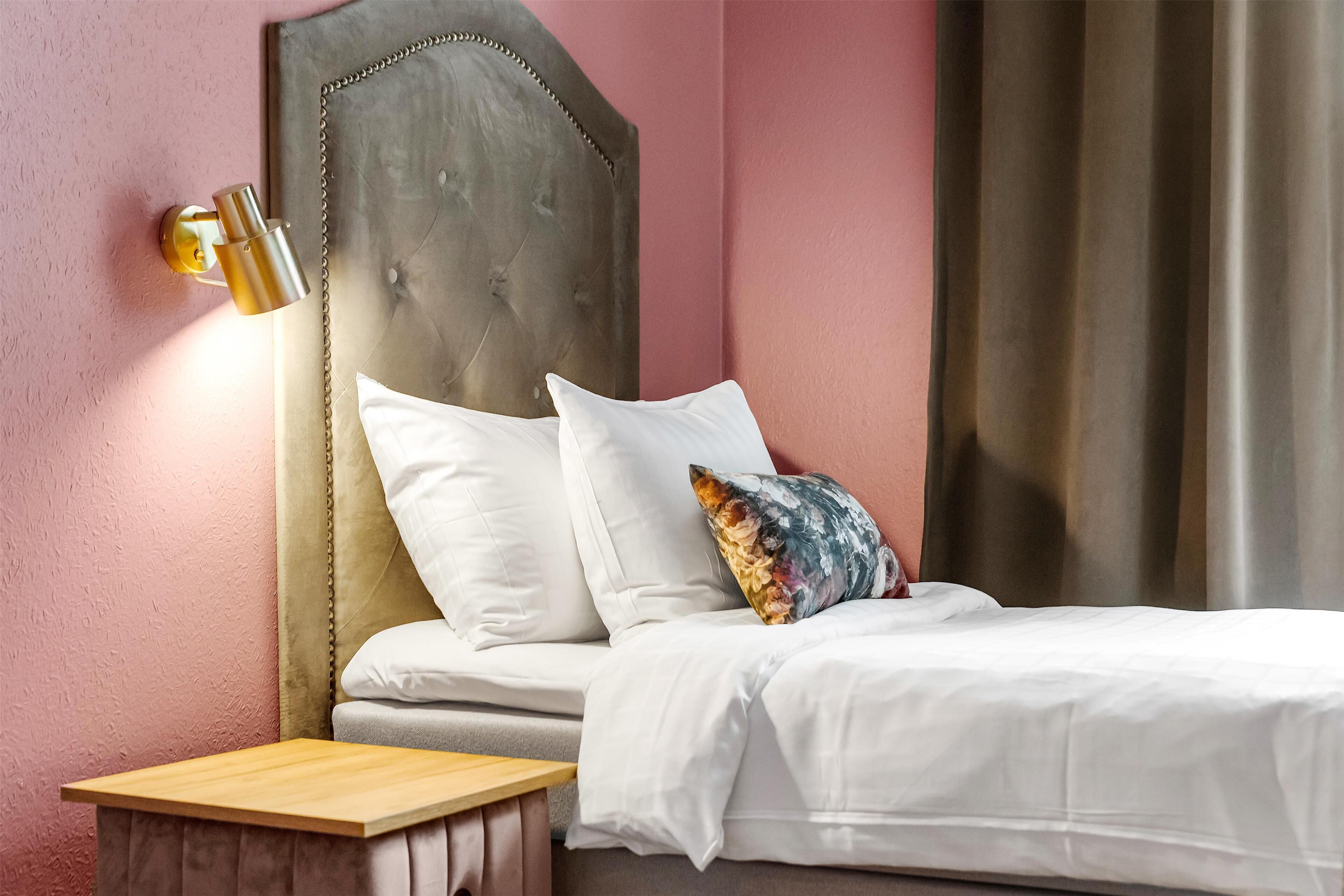 A bedroom with a bed, pillows, a nightstand, a wall-mounted lamp, and a curtain.