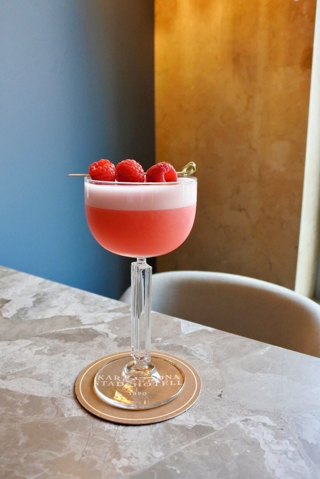 A cocktail in a coupe glass garnished with raspberries on a marble table with a coaster bearing the text "KARLSONA STADIOTEL 1890".