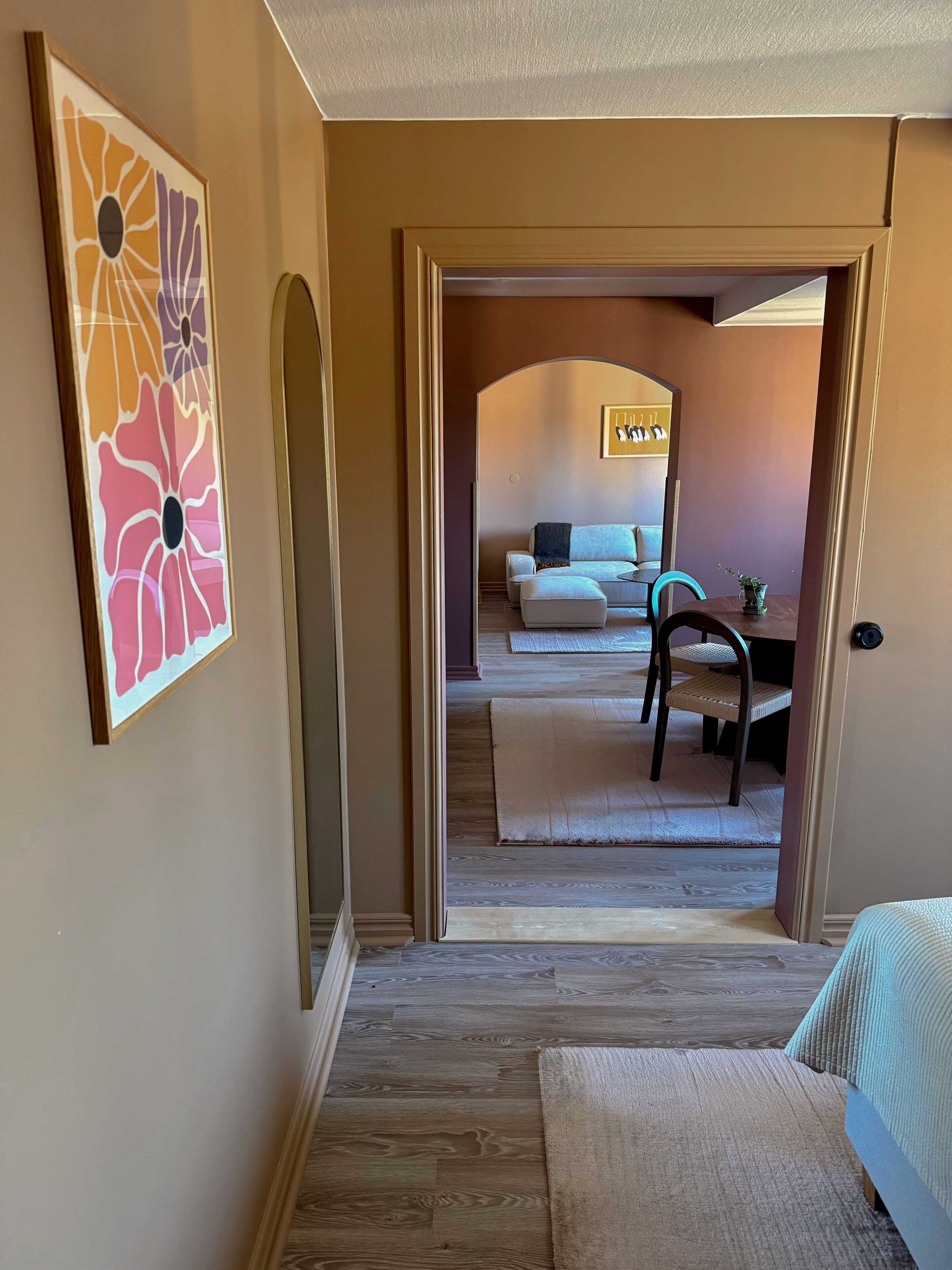 Ein Flur, der in ein Wohnzimmer mit Sofa, Couchtisch und Sessel führt. An der Wand hängt ein gerahmtes Kunstwerk und rechts befindet sich eine Tür.
