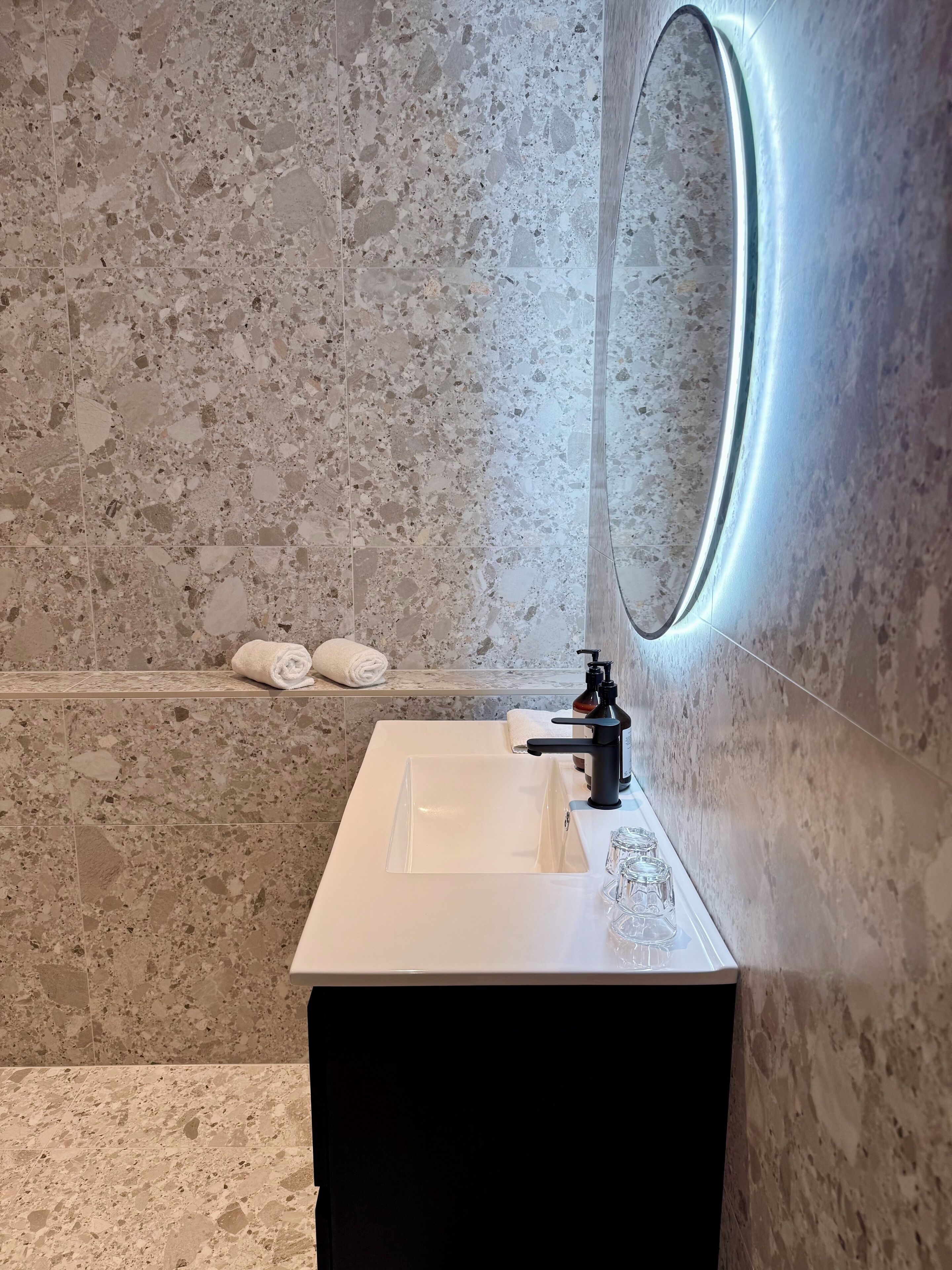 Un baño con lavamanos blanco, grifo negro y espejo con marco iluminado. Hay toallas en la pared y una botella de jabón en el lavamanos.