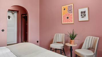 A bedroom with a bed, two chairs, a small table with a plant, and two framed artworks on the wall.
