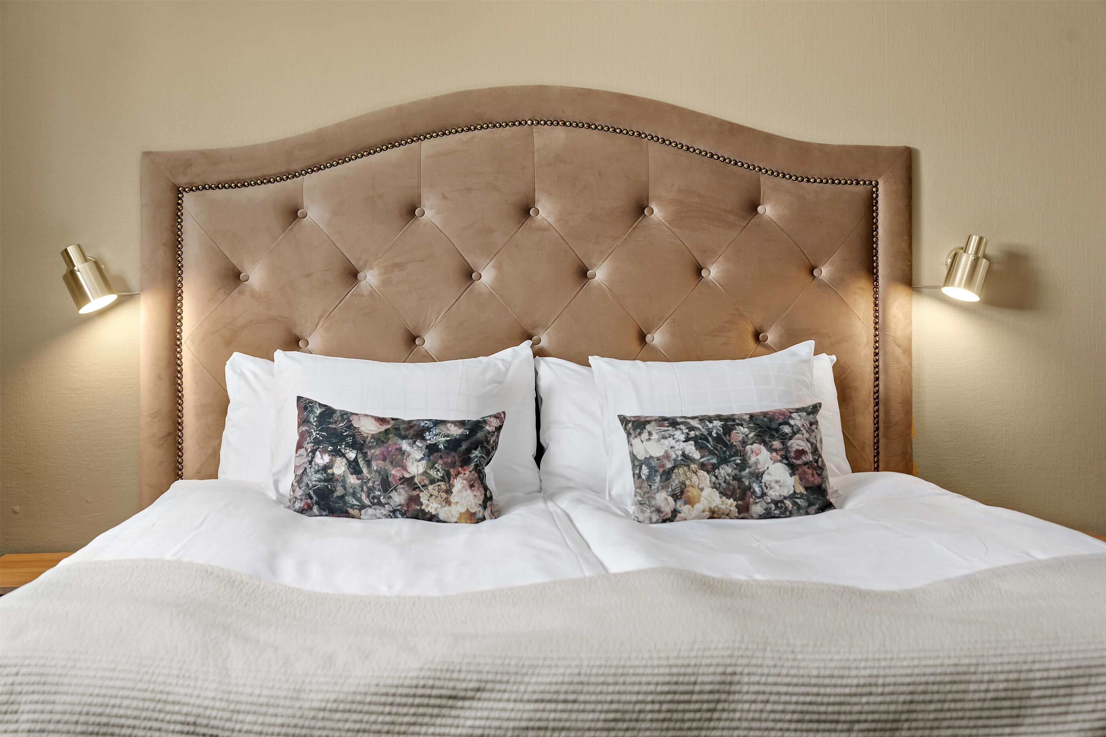 Una cama con cabecera tapizada, dos almohadas blancas y dos almohadas con estampado floral.