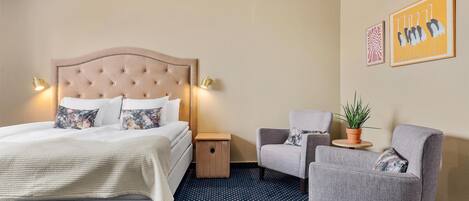 A hotel room with a large bed, two bedside tables, a grey armchair, a small side table with a plant, and two framed artworks on the wall.