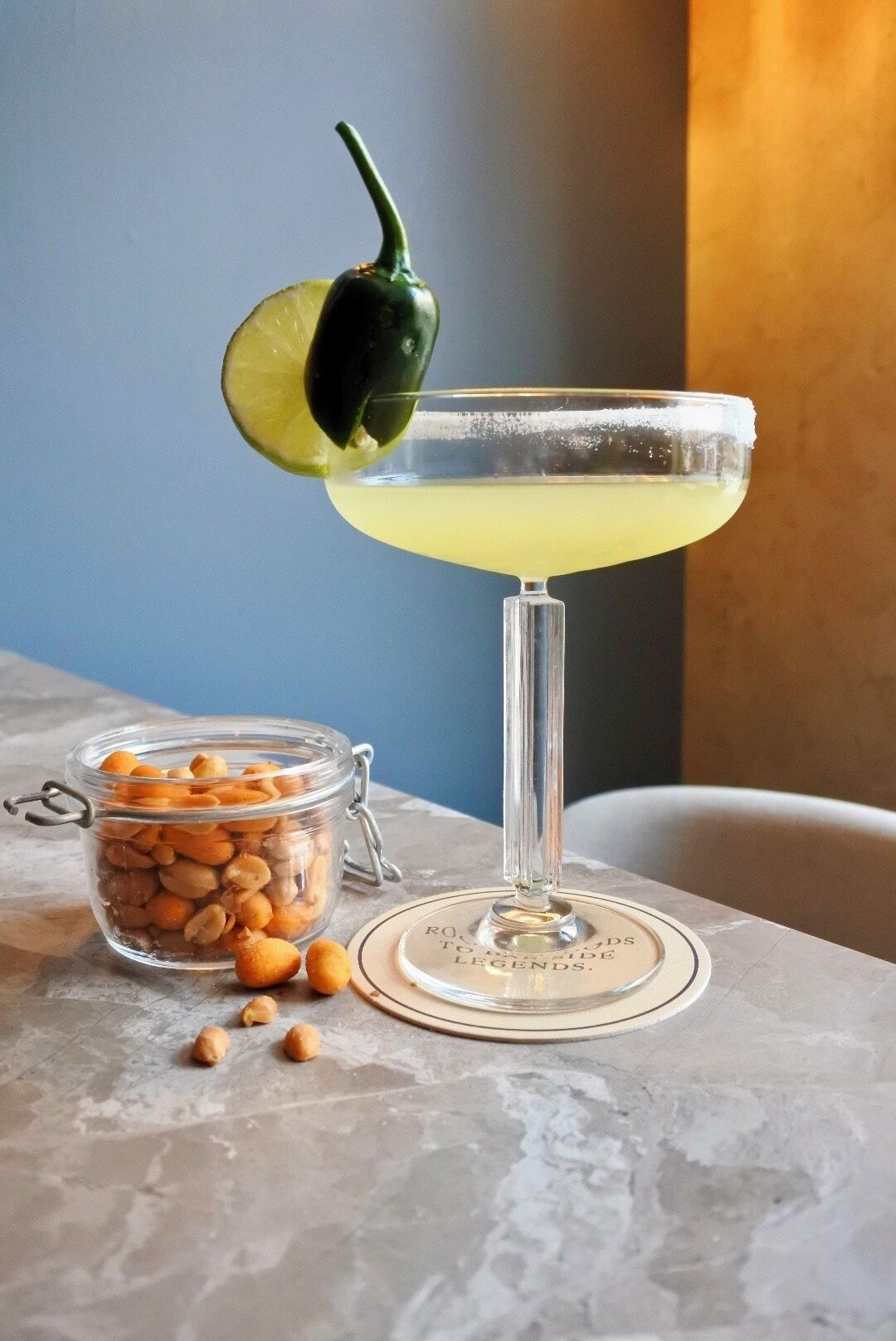 A martini glass with a lime garnish on a marble table, next to a jar of peanuts and a small plate with a quote.