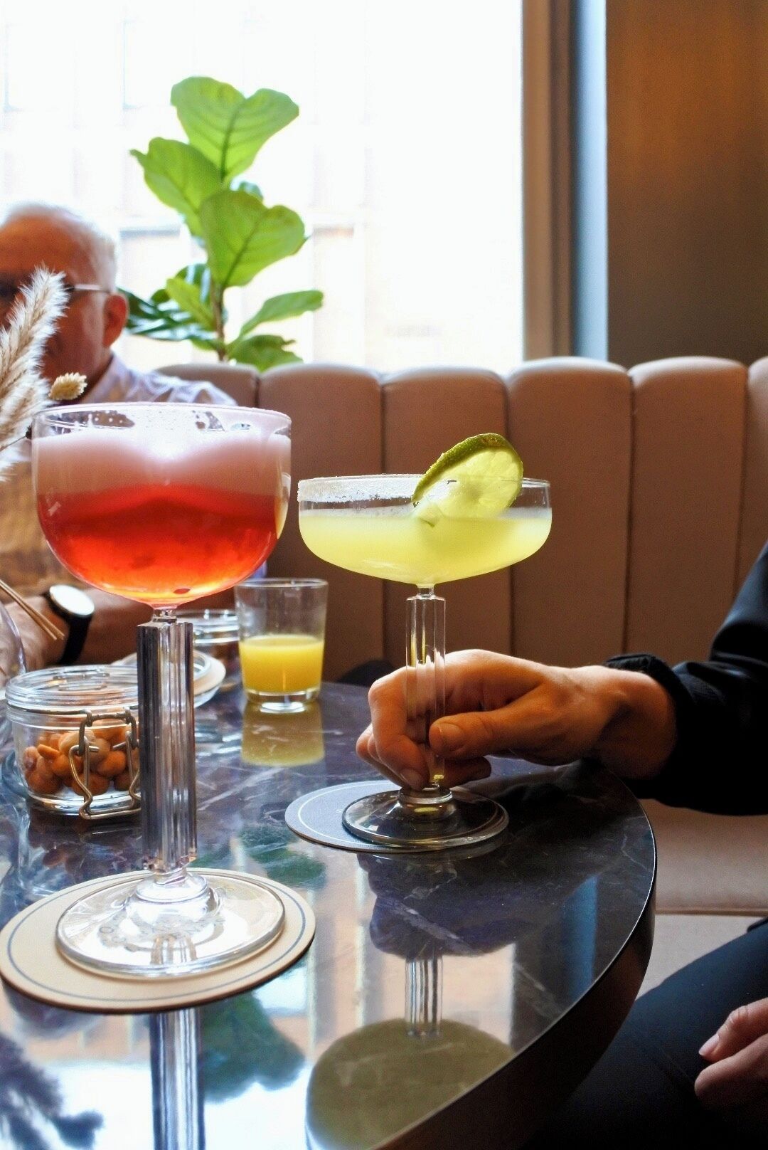Una mesa con dos cócteles, un vaso de jugo de naranja y un recipiente con nueces.