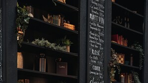 Un interior acogedor con un sofá de color oscuro decorado con cojines estampados, una estantería llena de libros y adornos, y un menú colgado en la pared que muestra bebidas.