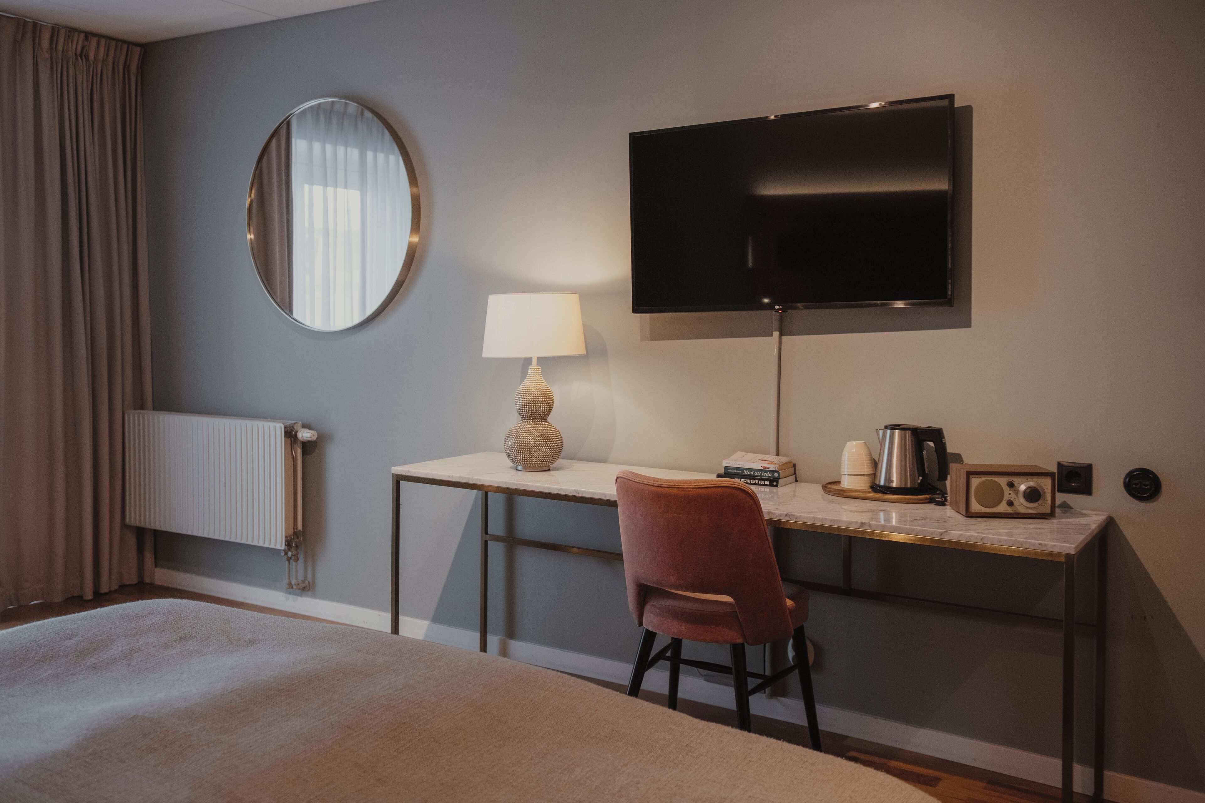 A hotel room with a bed, a desk with a lamp, a TV, a radio, and a radiator.