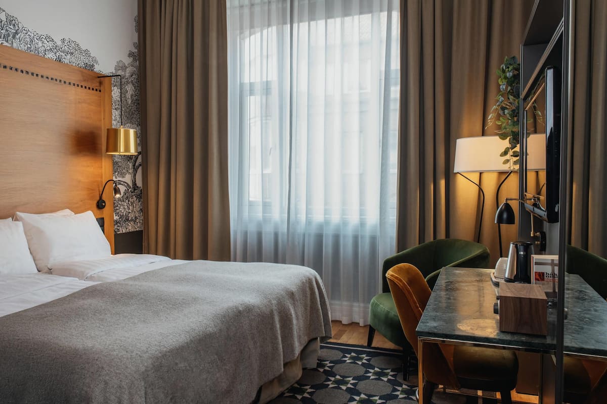 A hotel room with a bed, a desk, a chair, a lamp, and a window with curtains.