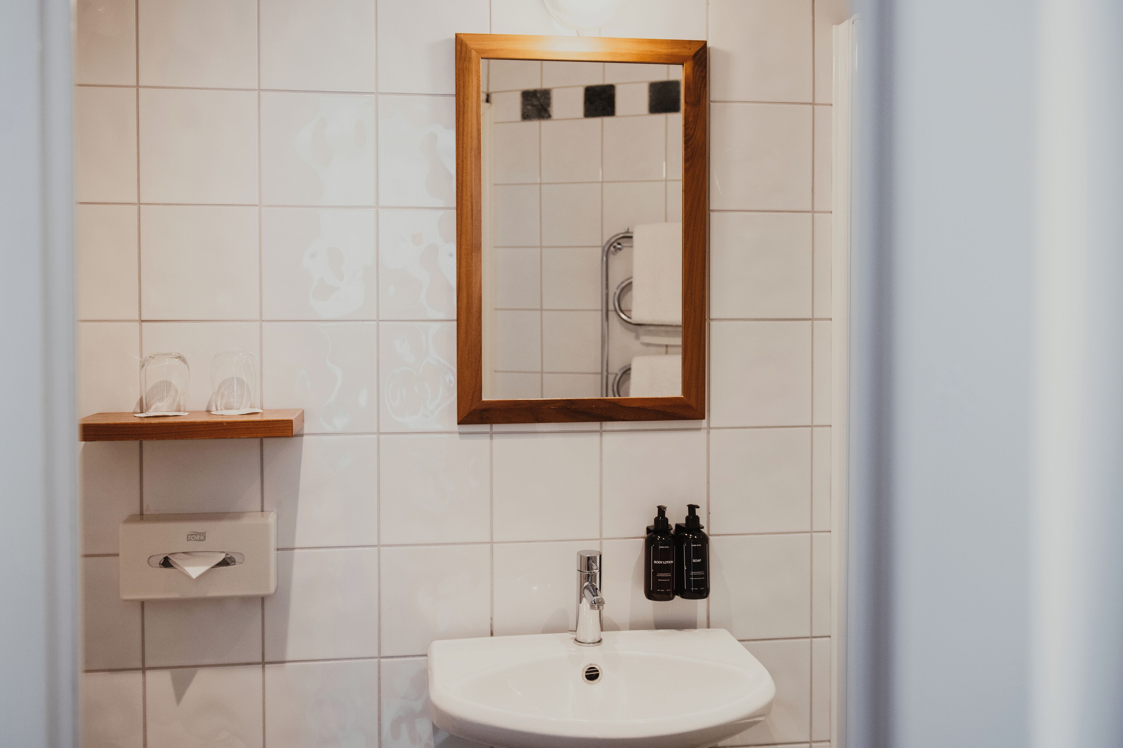 A bathroom with a white sink, a wooden-framed mirror, a towel rack, and two black bottles on the wall.