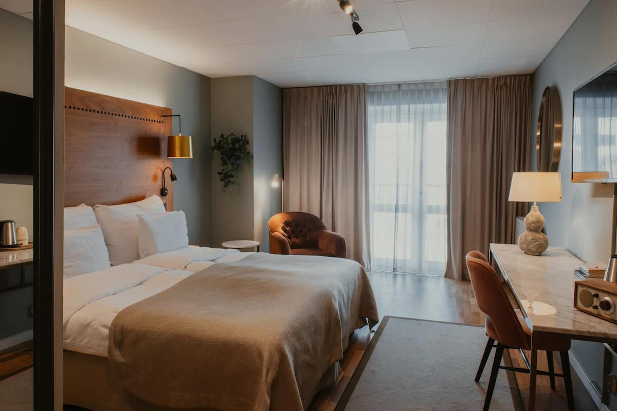 A hotel room with a large bed, a desk with a chair, a plant, and a window with curtains.