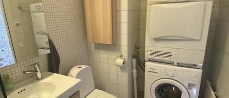 Et lille badeværelse med vask, toilet og stablet vaskemaskine og tørretumbler.