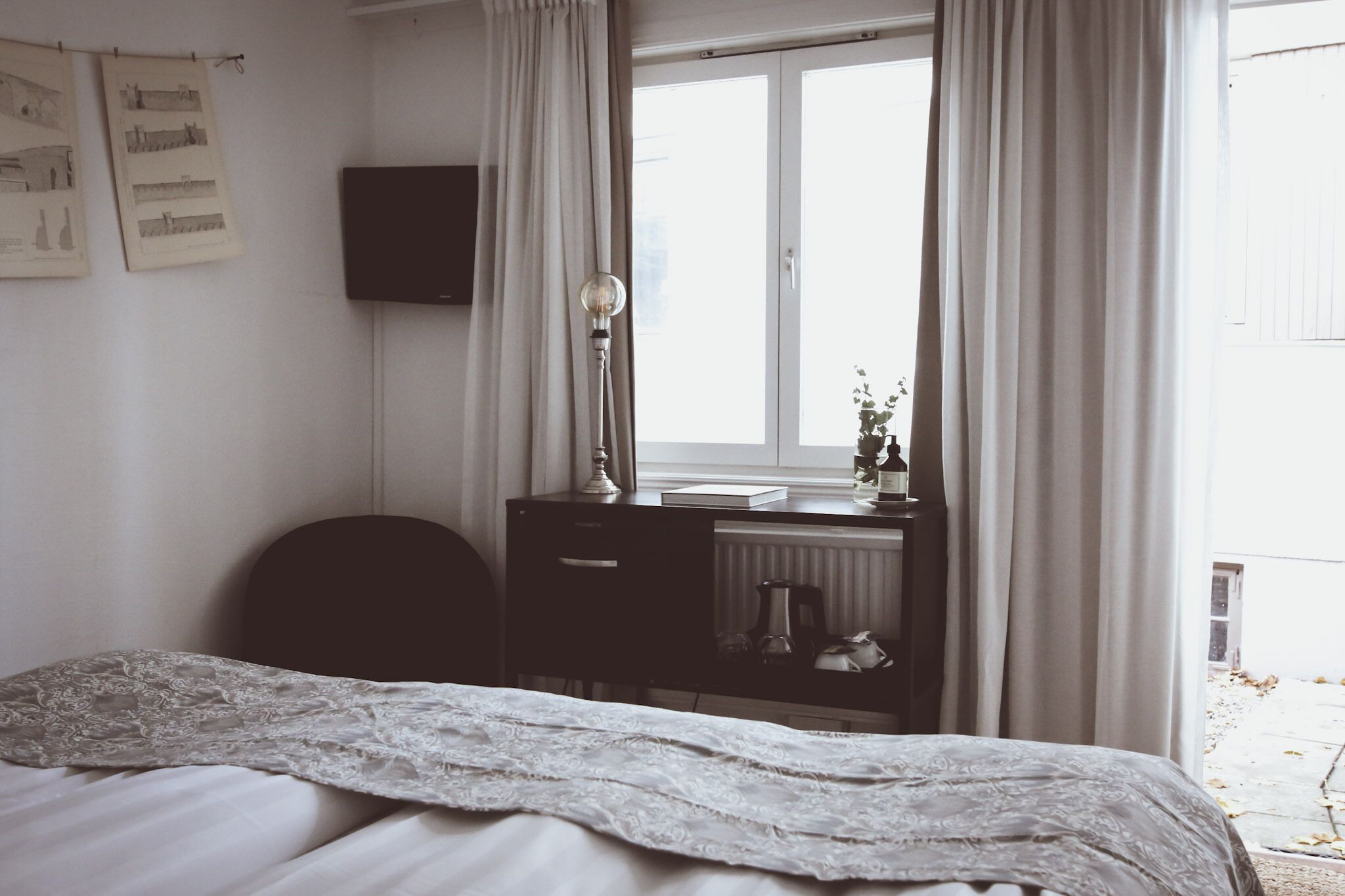 Une chambre à coucher avec un lit, un bureau avec une lampe, un radiateur et une fenêtre avec des rideaux.