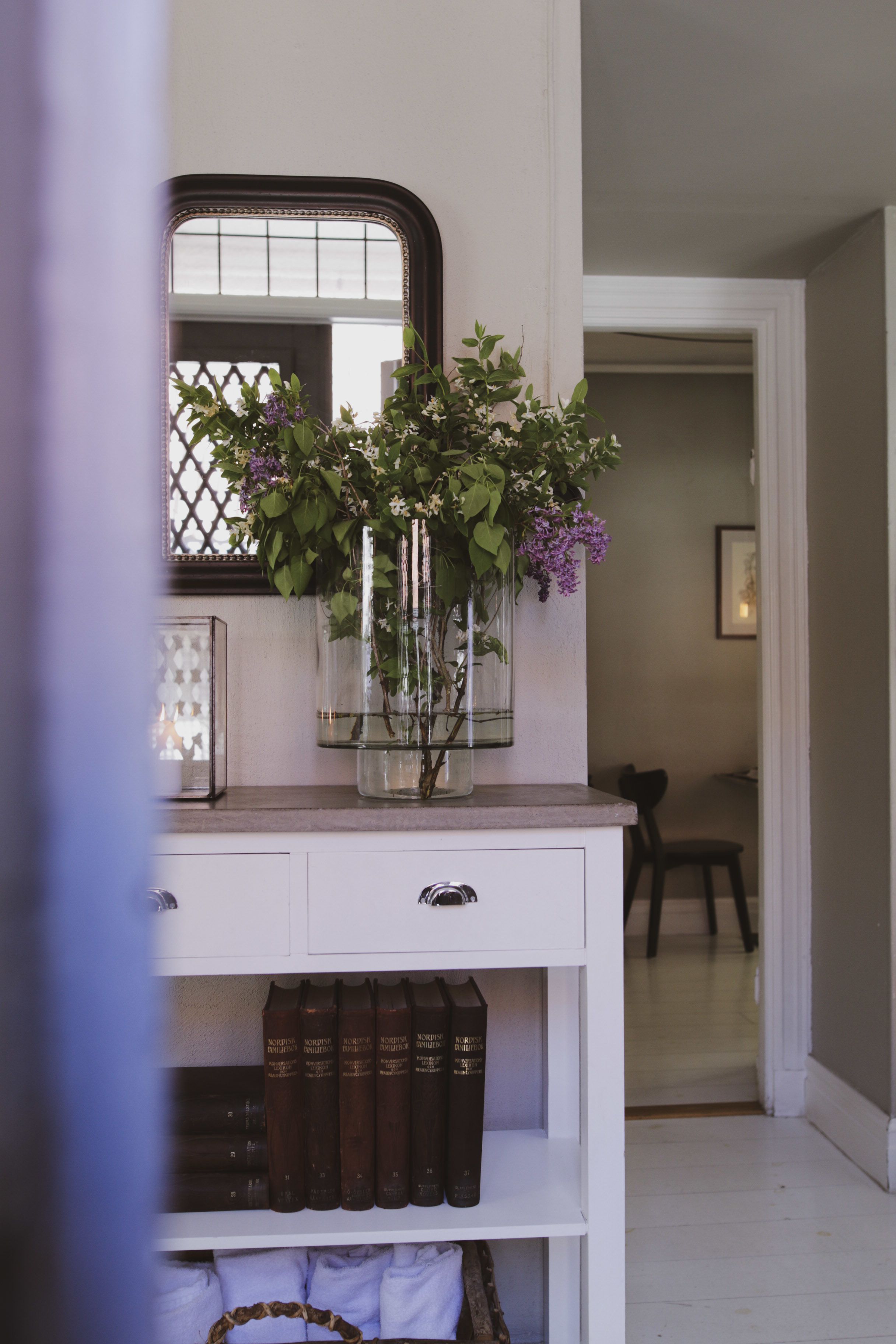 Una habitación con una consola blanca, un espejo y un florero con flores. Hay libros en el estante inferior y una silla al fondo.