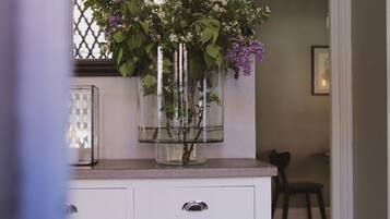 Una habitación con una consola blanca, un espejo y un florero con flores. Hay libros en el estante inferior y una silla al fondo.