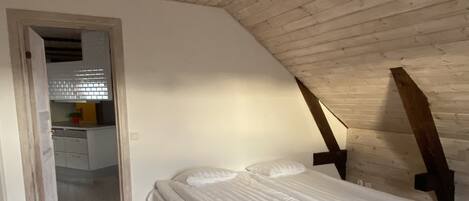 Un dormitorio con techo inclinado, una cama, piso de madera y vista a una cocina por una puerta abierta.