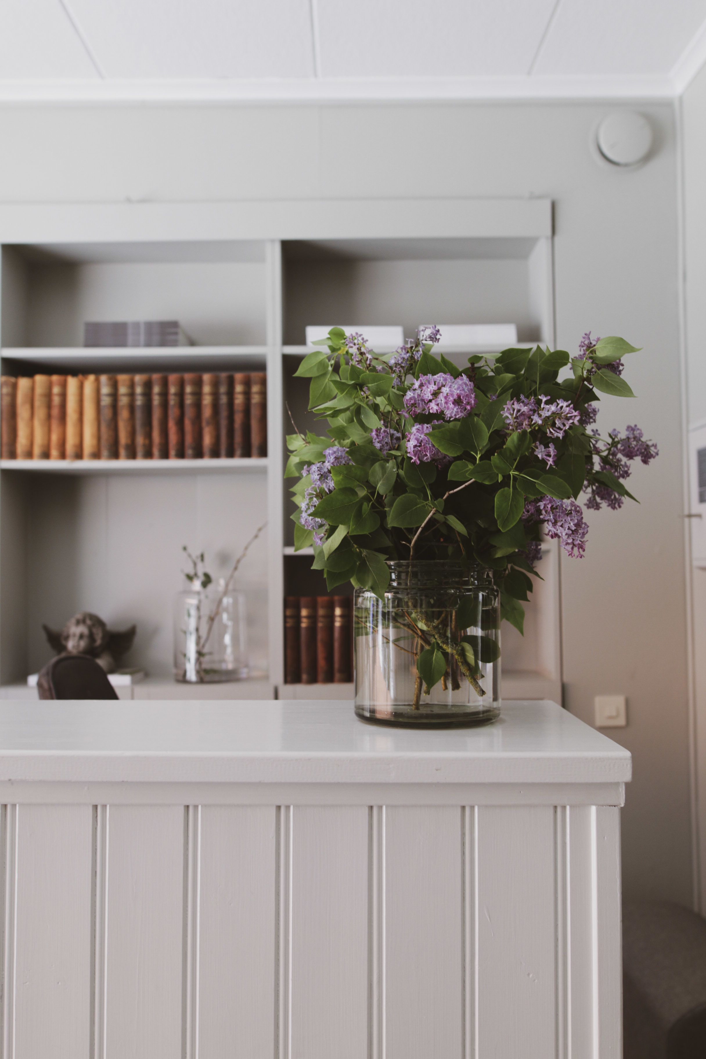 Una mesa blanca con un florero de vidrio con flores moradas. Detrás de la mesa, hay una estantería con libros y un objeto decorativo.