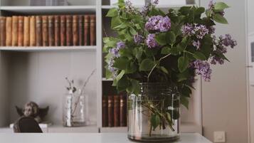 Una mesa blanca con un florero de vidrio con flores moradas. Detrás de la mesa, hay una estantería con libros y un objeto decorativo.