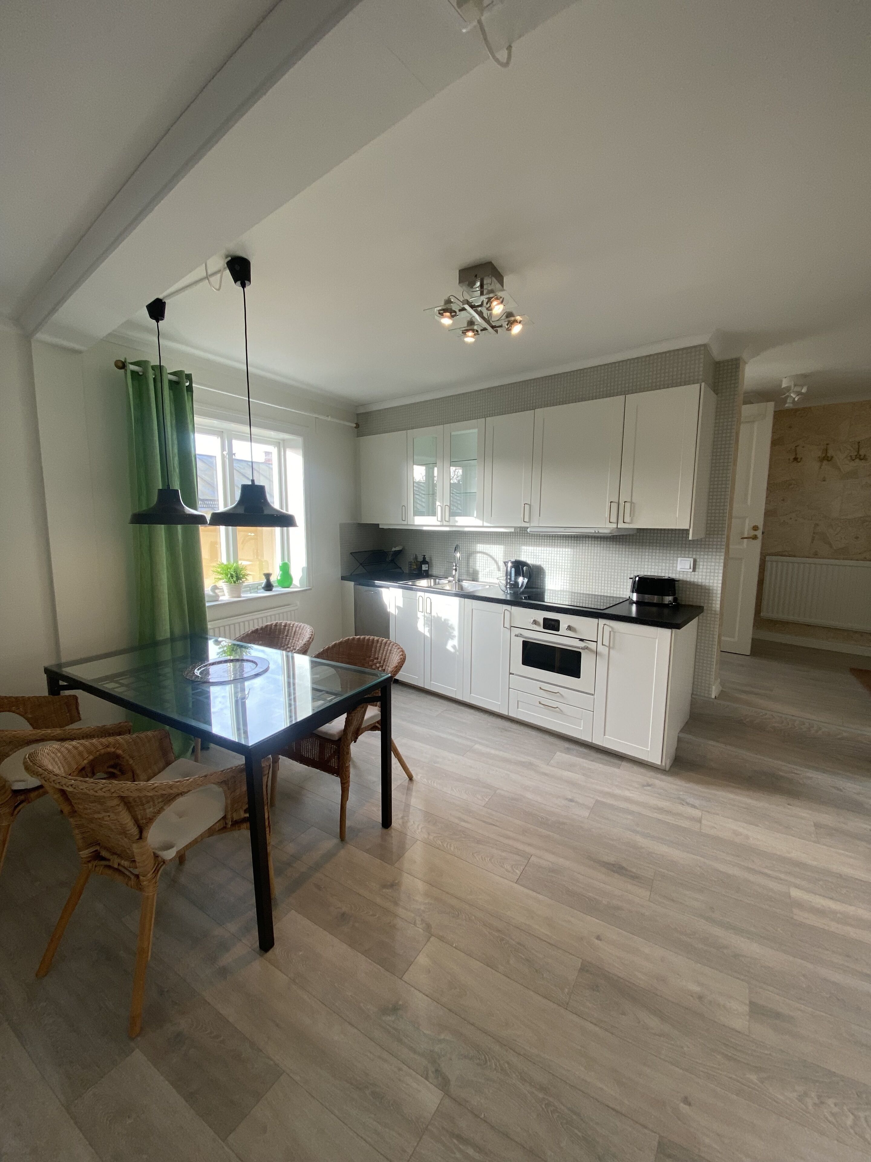 A modern kitchen with white cabinets, a glass dining table, wicker chairs, and a wooden floor.