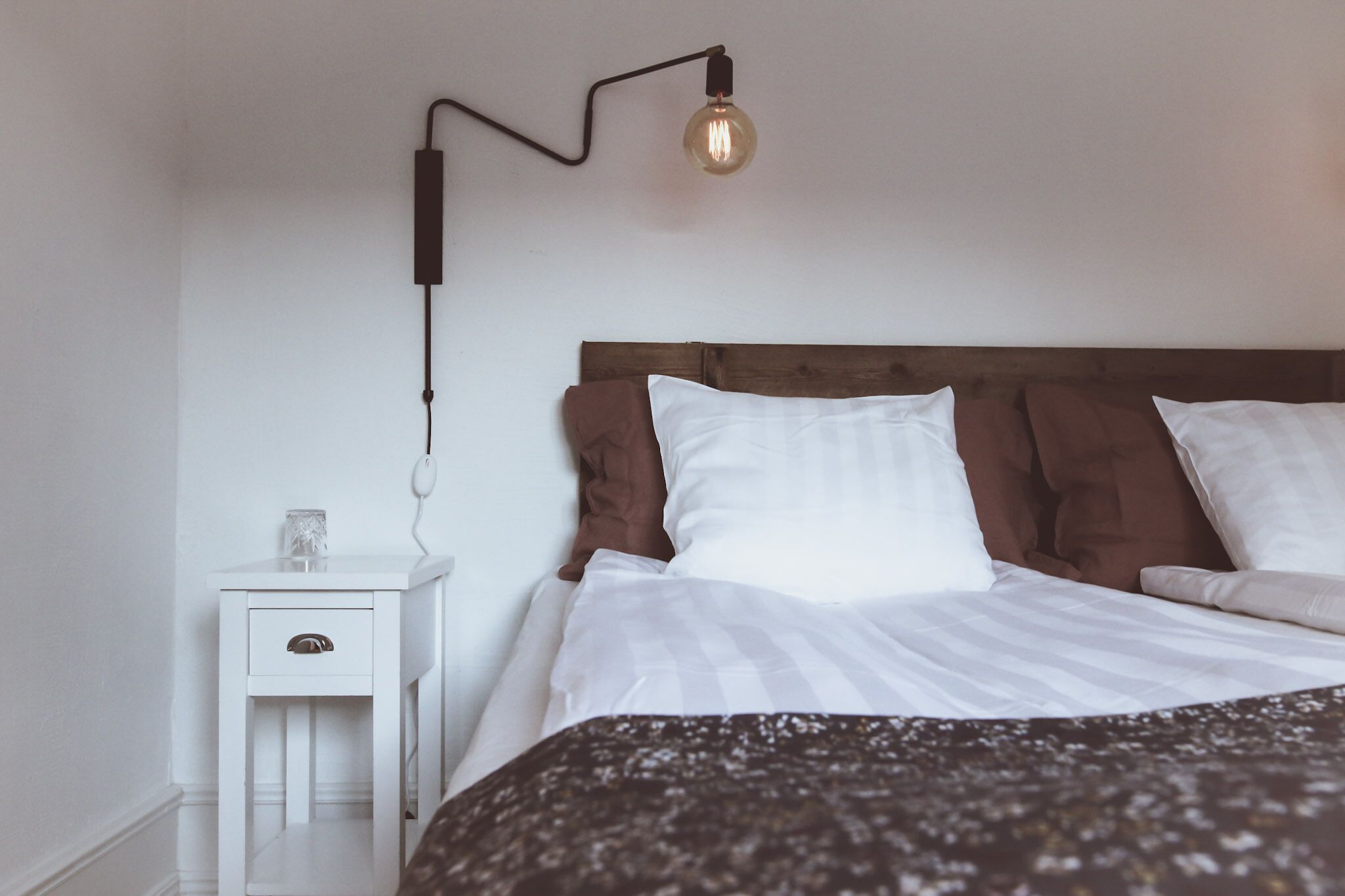 A neatly made bed with white and brown bedding, a small bedside table with a drawer, and a wall-mounted light fixture.