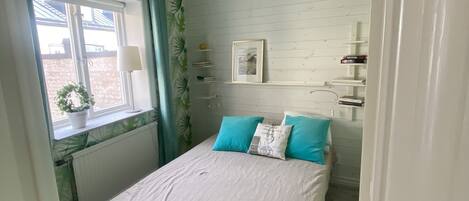 A bedroom with a bed, a window with curtains, a radiator, a ceiling fan, and a wall-mounted shelf with books.