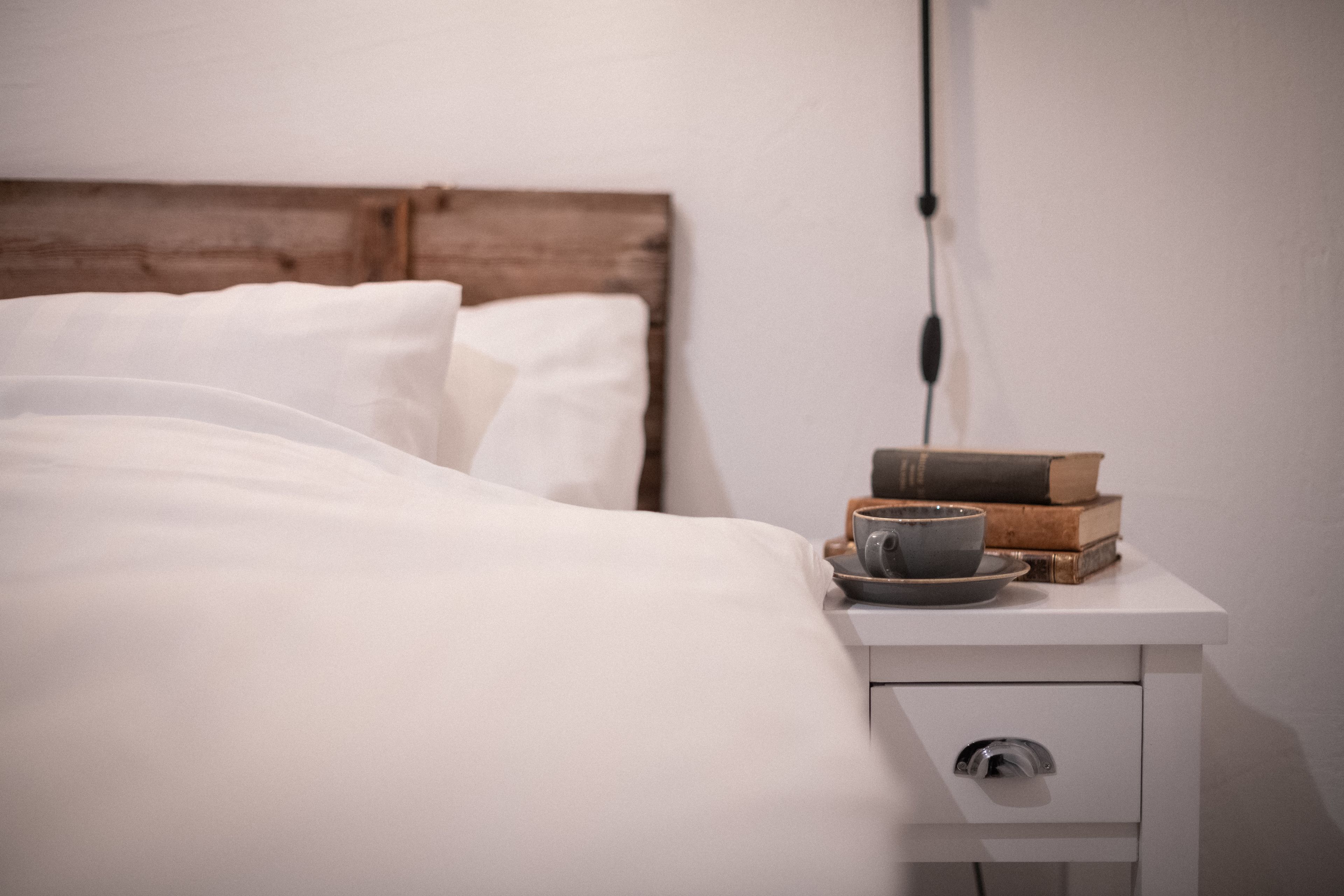A neatly made bed with white linens, a bedside table with a cup and books, and a wall socket.
