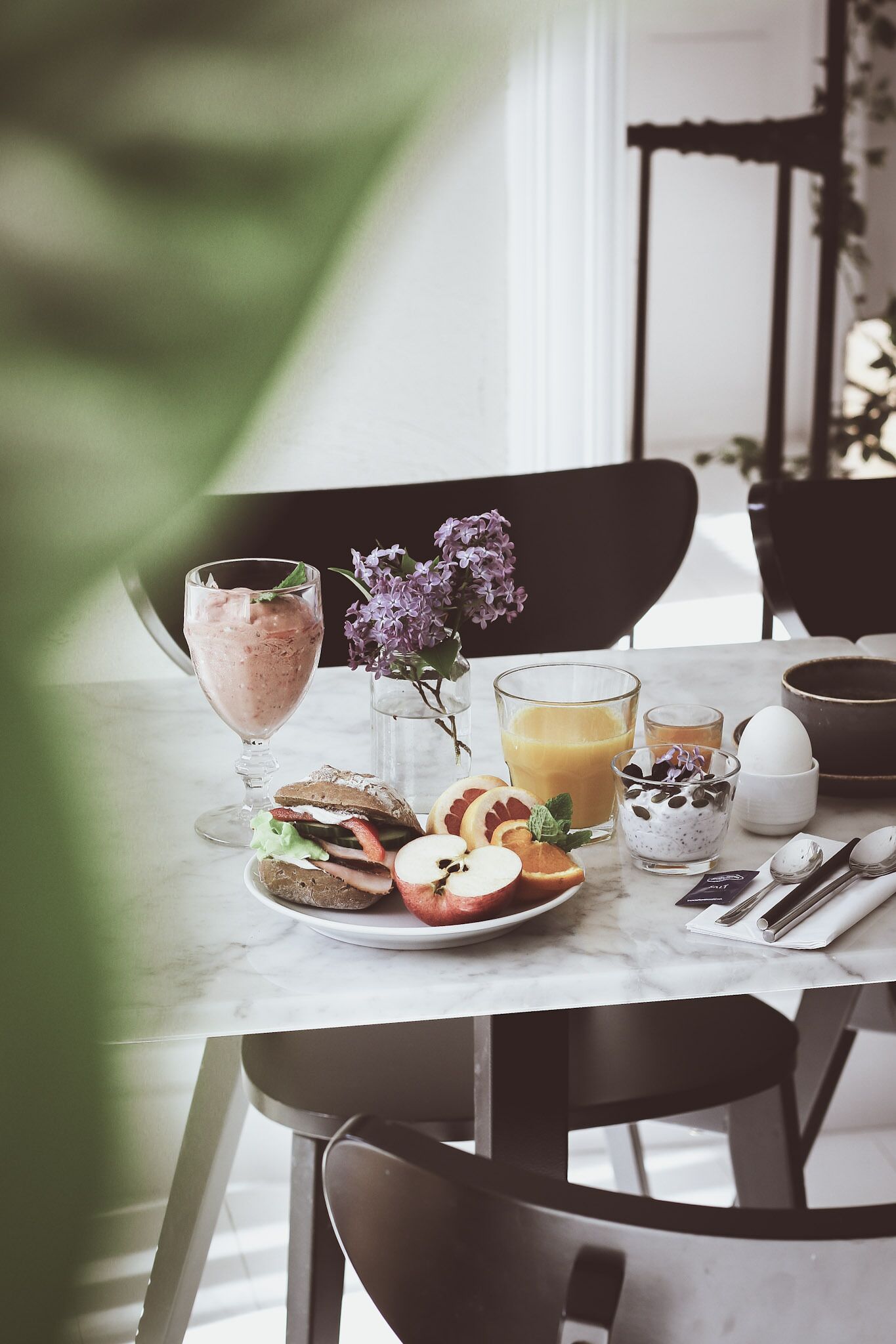A breakfast table with a glass of smoothie, a glass of orange juice, a bowl of yogurt, a plate of toast with fruit, and a vase with flowers.
