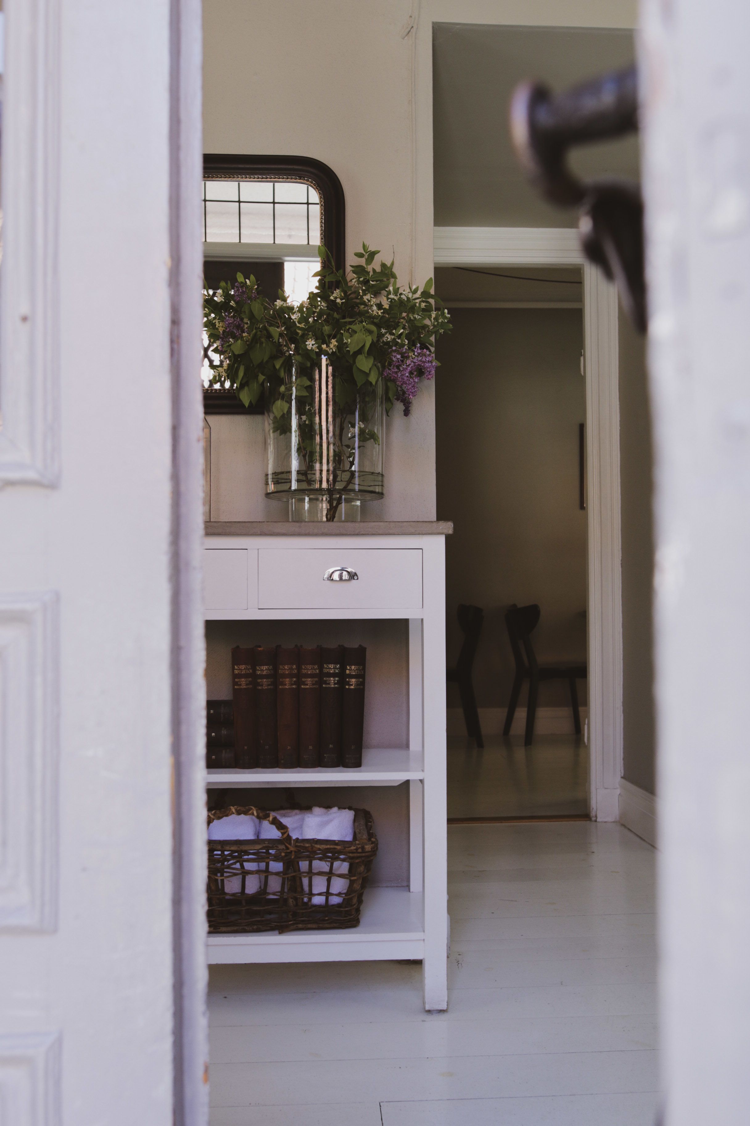 A white console table with a mirror, a vase with flowers, and bookshelves.