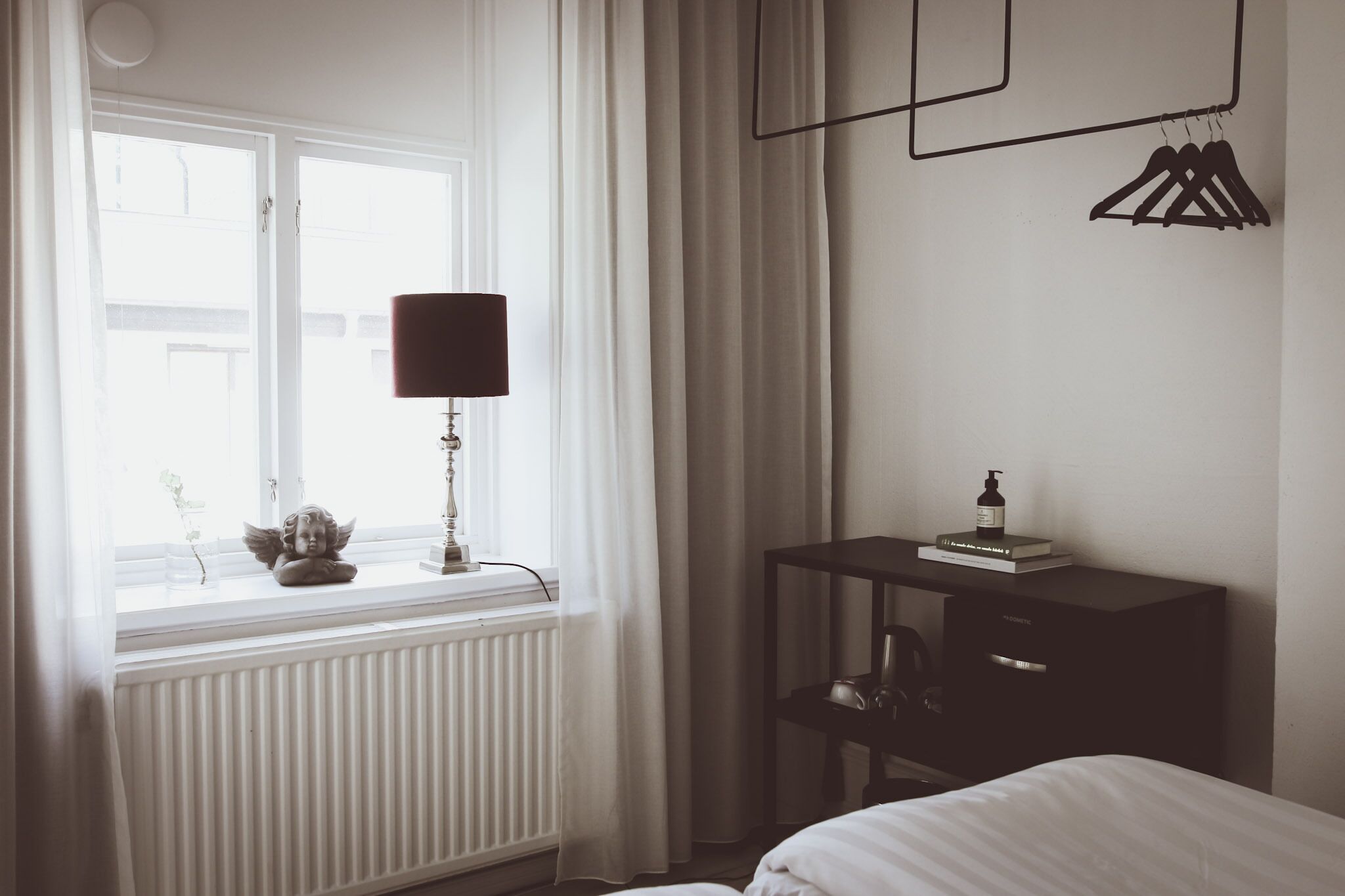 Quarto com cama, mesa de cabeceira com luminária, radiador e janela com cortinas.