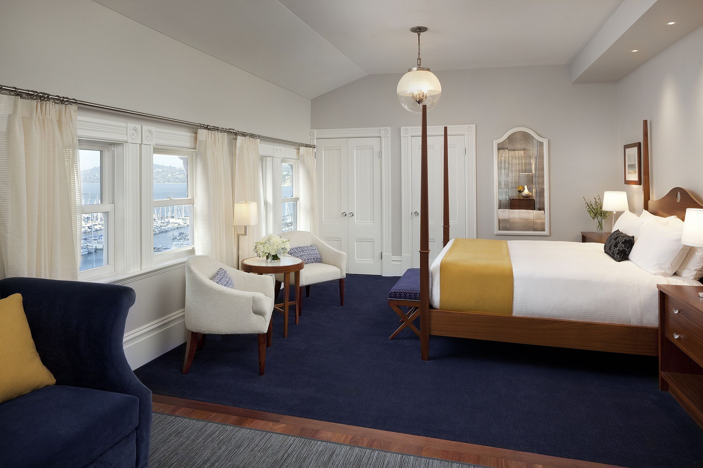 A hotel room with a large bed, a seating area with a chair and a small table, a mirror, and a view of the harbor through the windows.