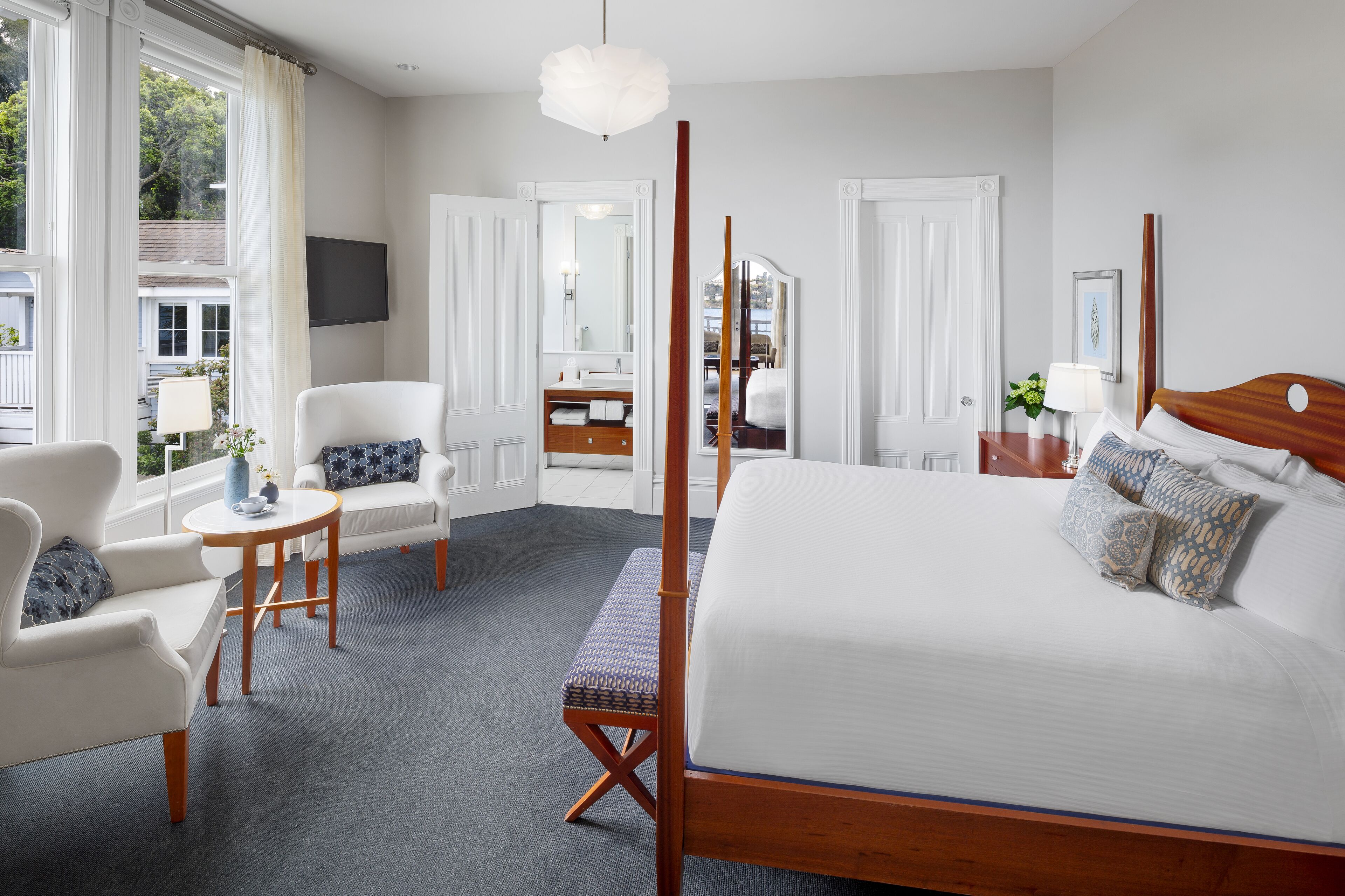 Una habitación de hotel moderna con una cama grande, una zona de estar con una silla y una mesita, un tocador con espejo, y vistas al exterior a través de amplios ventanales.
