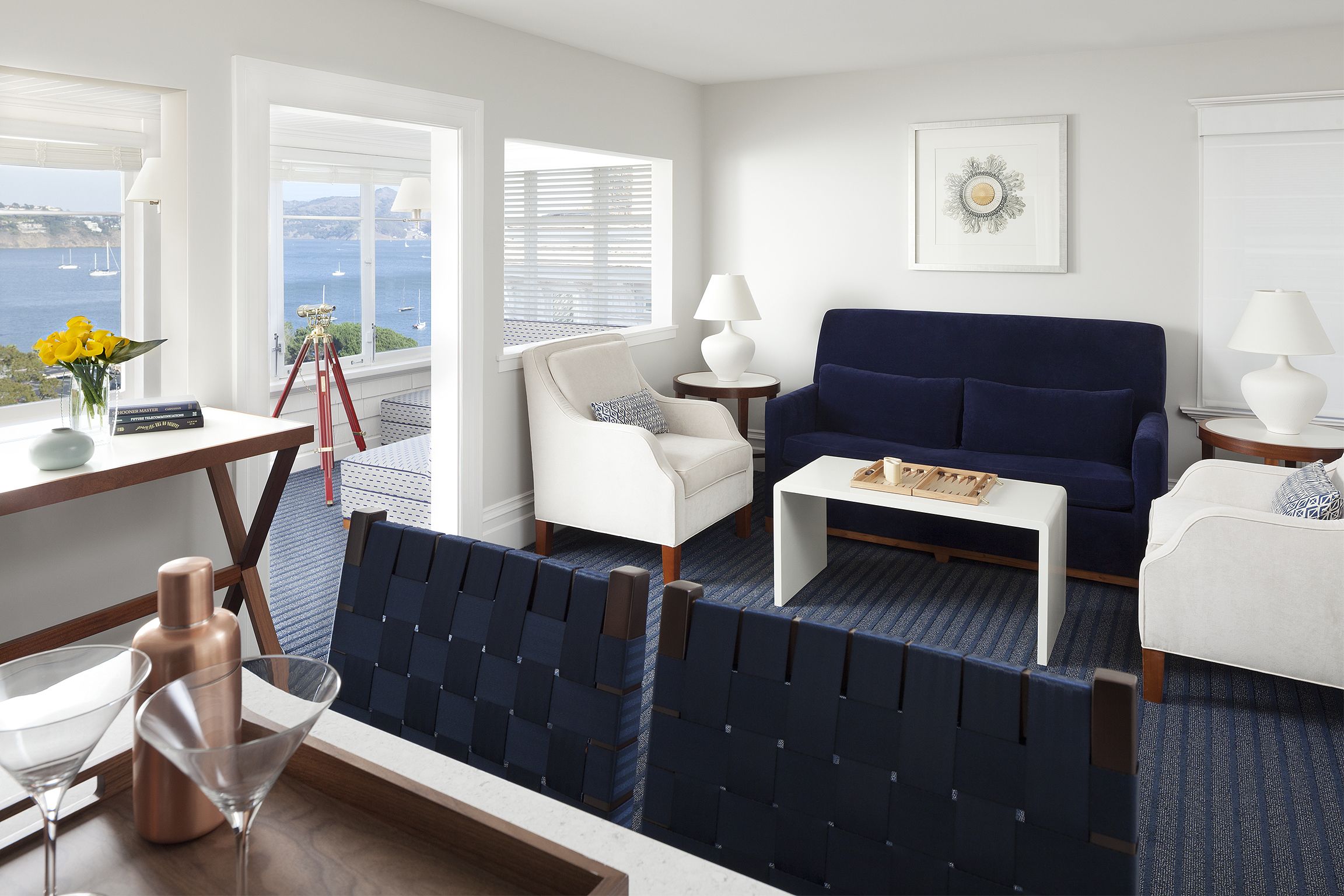 A modern living room with a sofa, armchairs, and a coffee table. There are two lamps, a framed artwork, and a view of the sea through large windows.