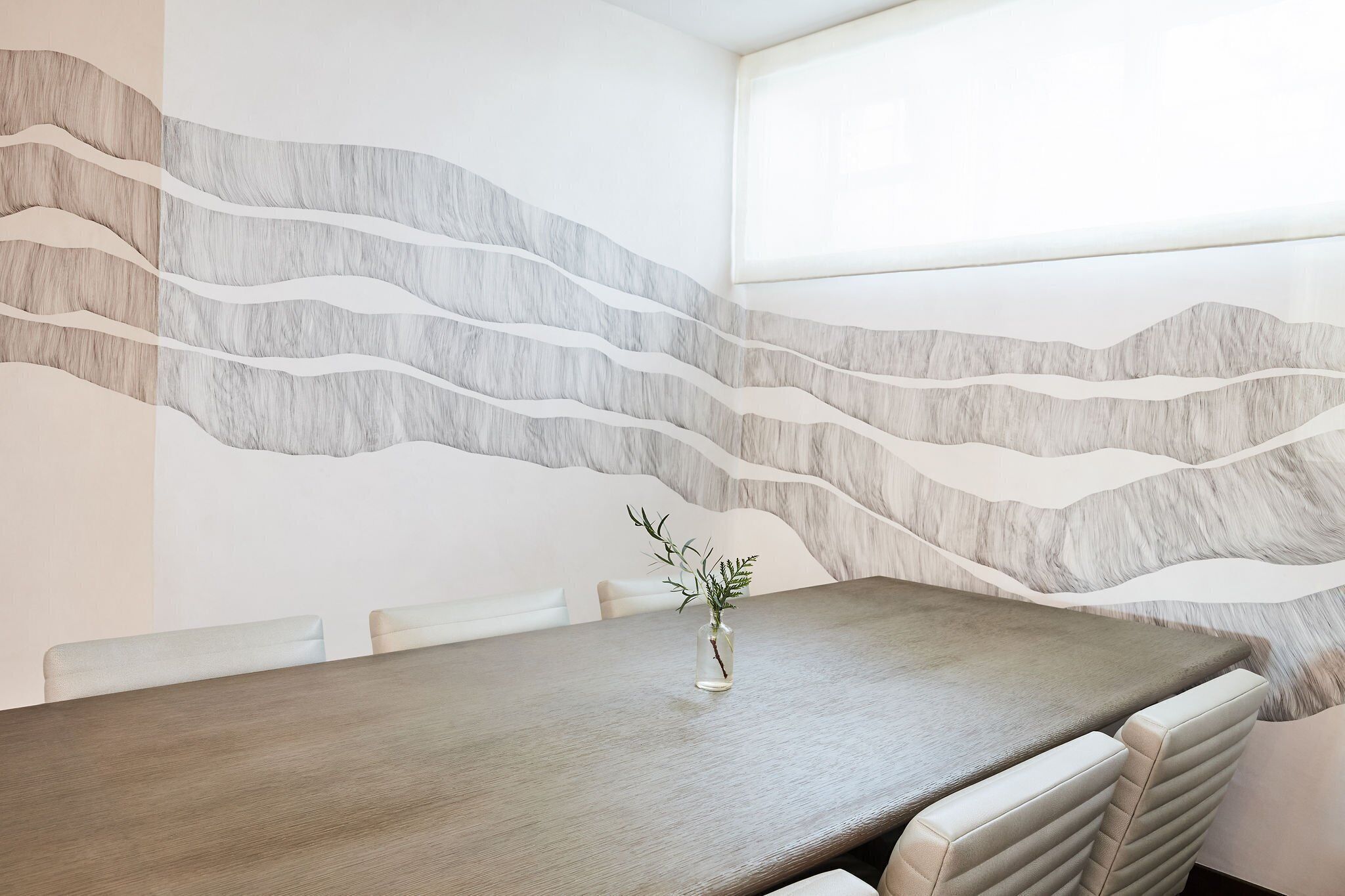 A conference room with a wooden table and beige upholstered chairs. A small vase with a plant is on the table. The wall features a wavy, textured design.