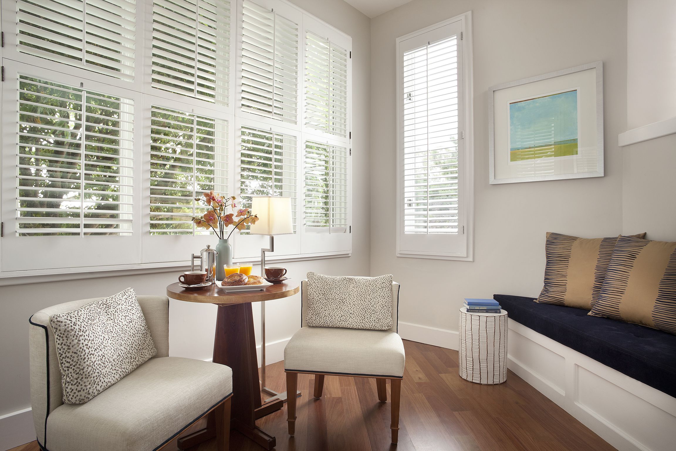 A room with a wooden floor, a round table with a lamp, two chairs, a sofa, and a window with blinds.