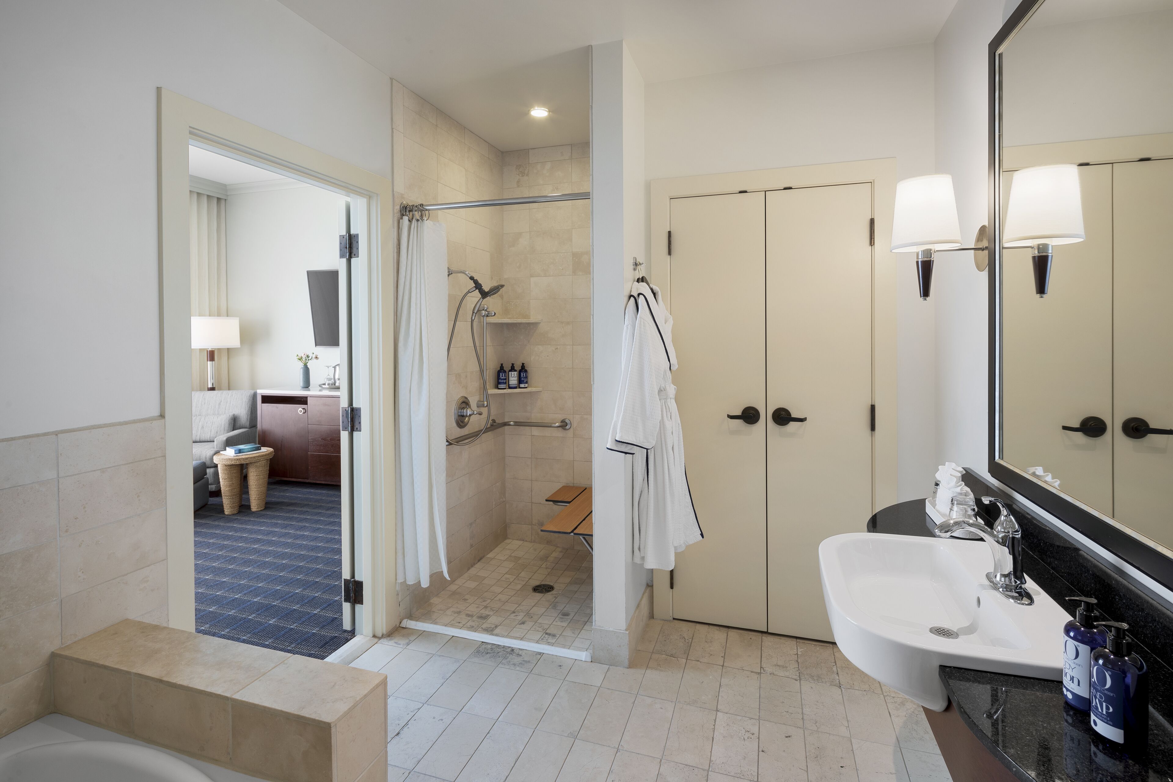 Une salle de bain moderne avec une baignoire, un lavabo et une douche. On y trouve un miroir, un porte-serviettes et une porte donnant sur une chambre.