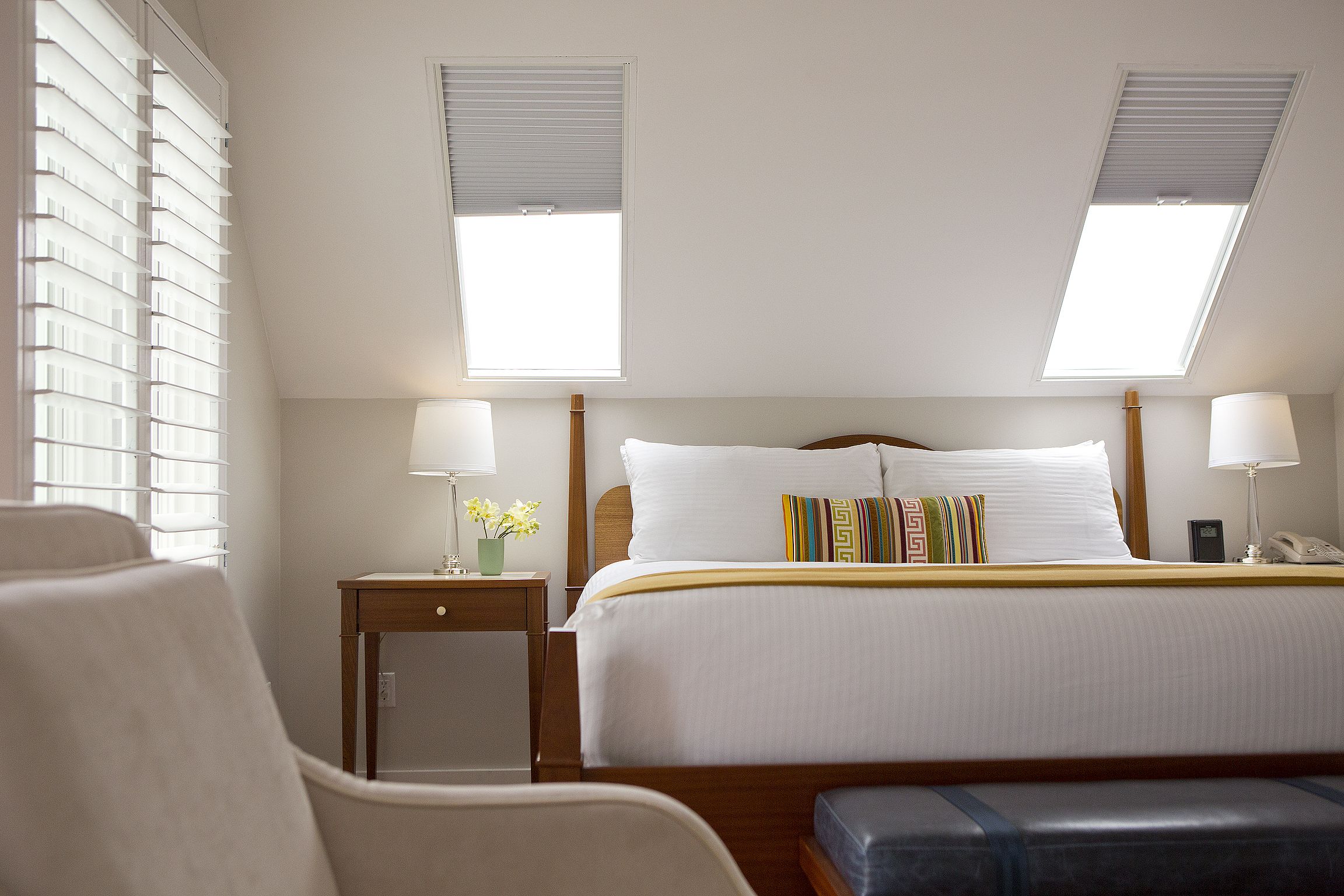 A hotel room with a large bed, two bedside lamps, a small bedside table with a lamp, a chair, and a window with blinds.