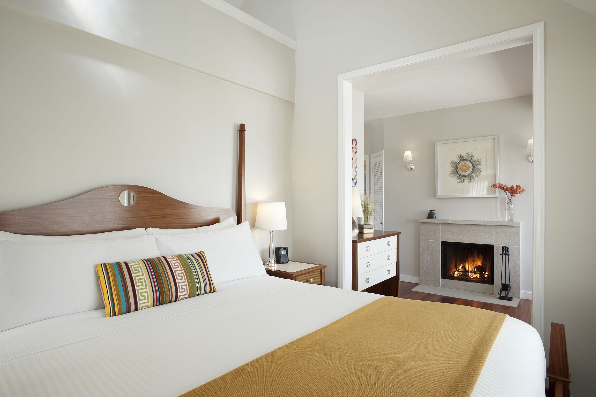 A hotel room with a large bed, a bedside table with a lamp, a nightstand, a fireplace, and a framed picture on the wall.