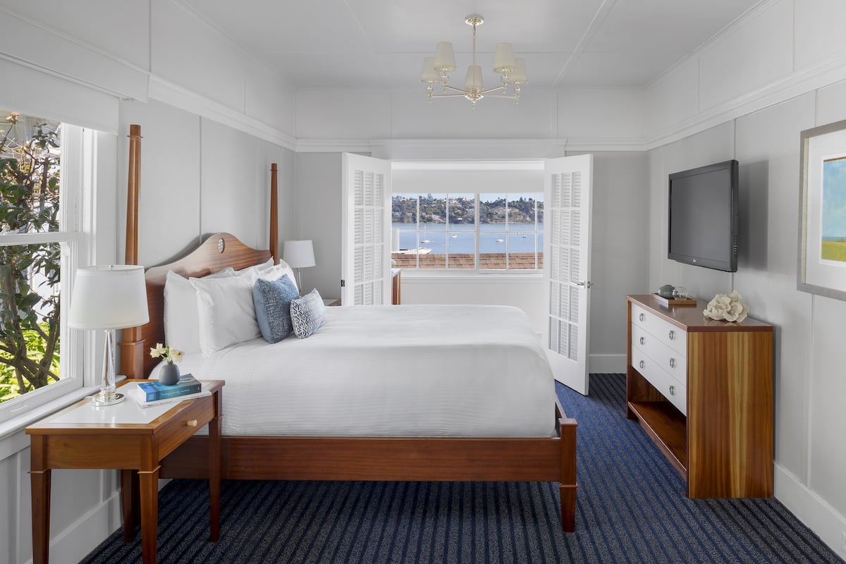 Ein Hotelzimmer mit einem großen Bett, einem Nachttisch mit Lampe, einem Schminktisch, einem Fernseher und einem Fenster mit Meerblick.