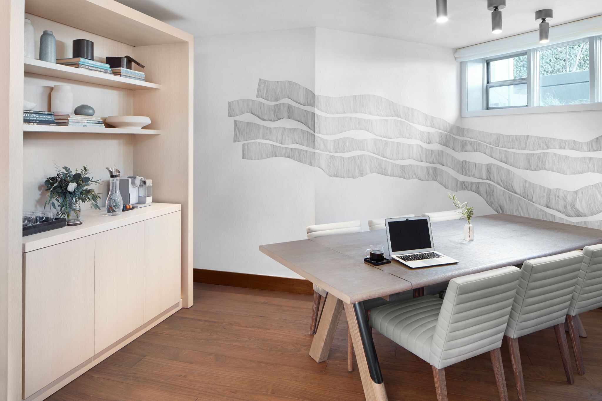 A modern dining area with a wooden table, white chairs, and a laptop. There are shelves with books and decorative items, and a wall with a wavy pattern.