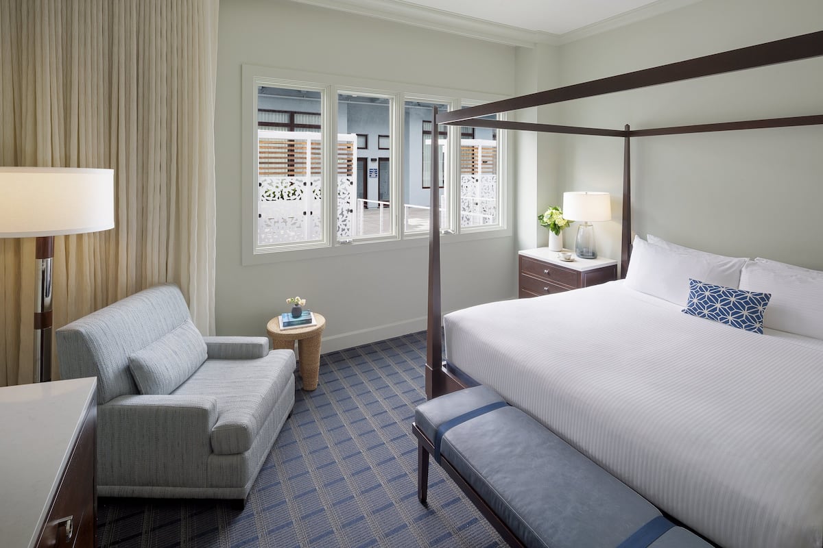 Ein Hotelzimmer mit Himmelbett, einer Couch, einem kleinen runden Tisch mit Lampe und Blick durch große Fenster nach draußen.