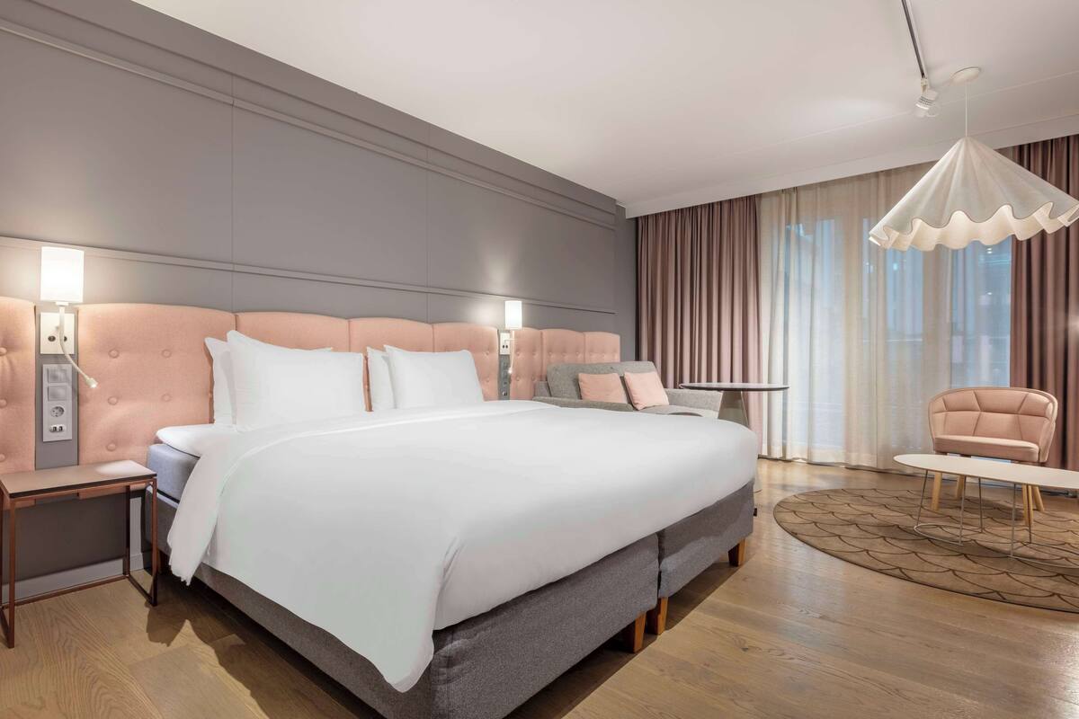 A modern hotel room with a large bed, bedside tables, a round coffee table, and a chandelier.