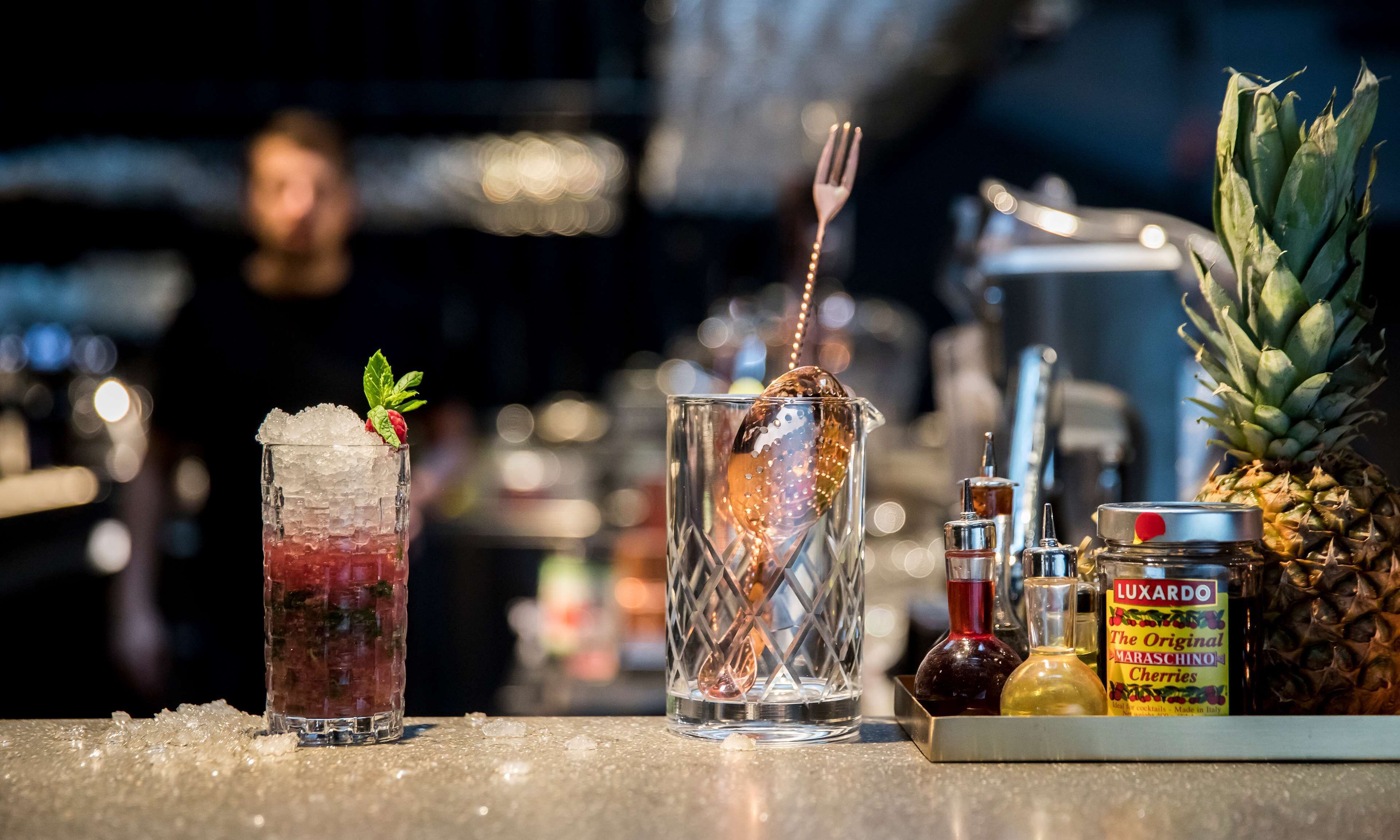 A bar setting with two cocktails, a pineapple, and a bottle of Luxardo cherries.