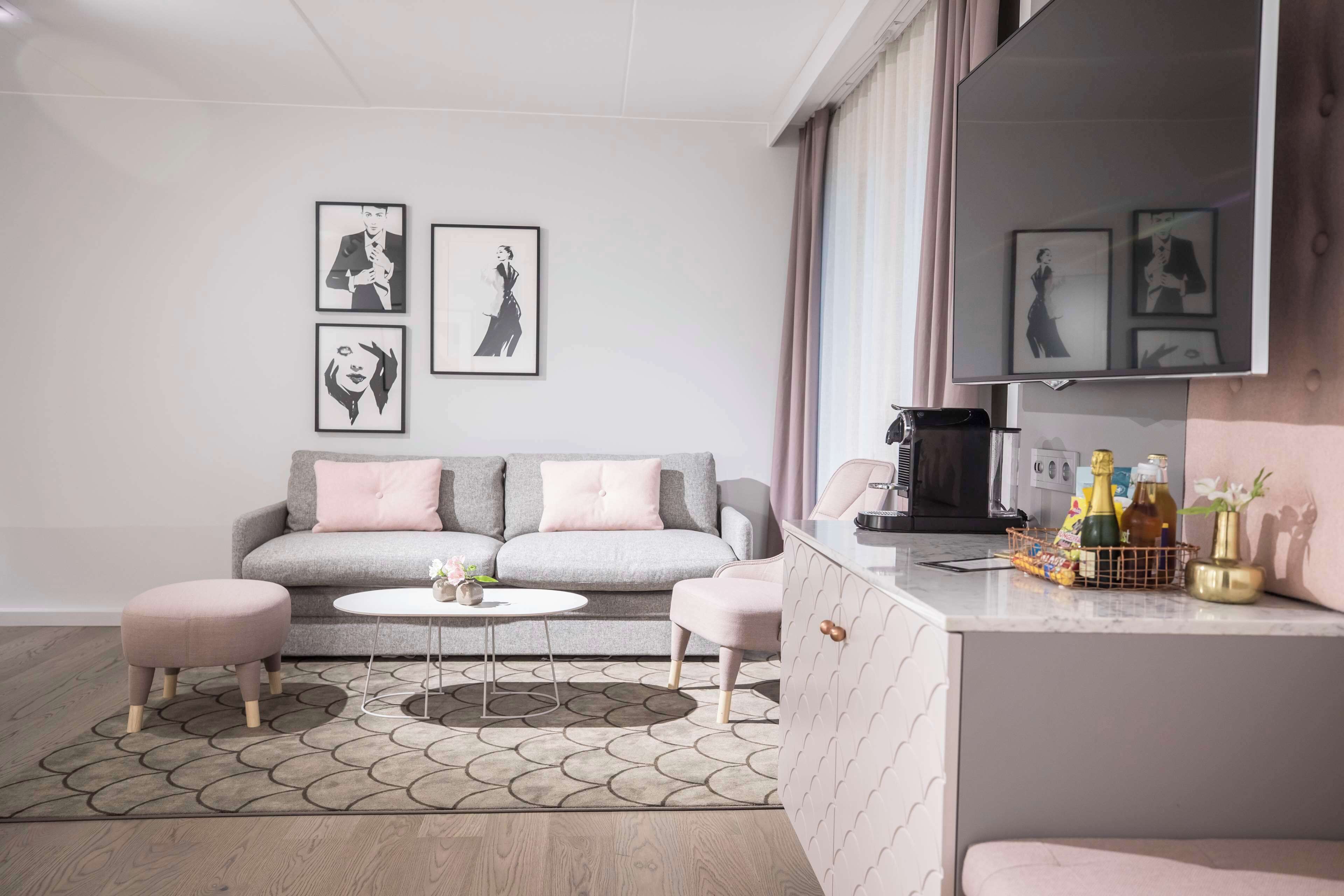 Una sala de estar moderna con un sofá gris, dos otomanas rosas, una mesa de centro redonda y una zona de cocina con encimera y armarios.