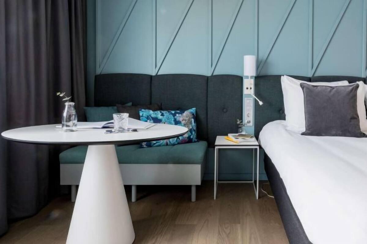 A modern hotel room with a bed, a round table, a small bedside table, and a wall-mounted power outlet.