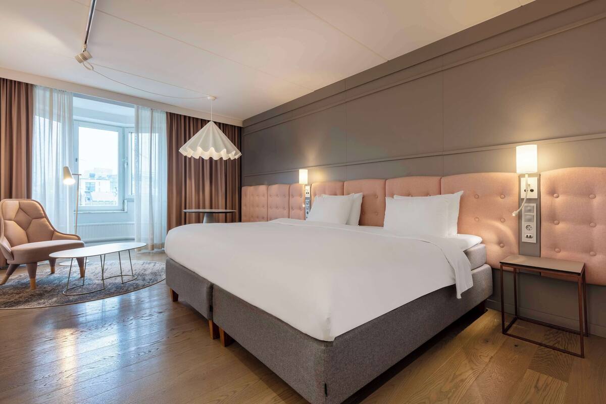 A modern hotel room with a large bed, a sitting area with a chair and table, and a view through large windows.