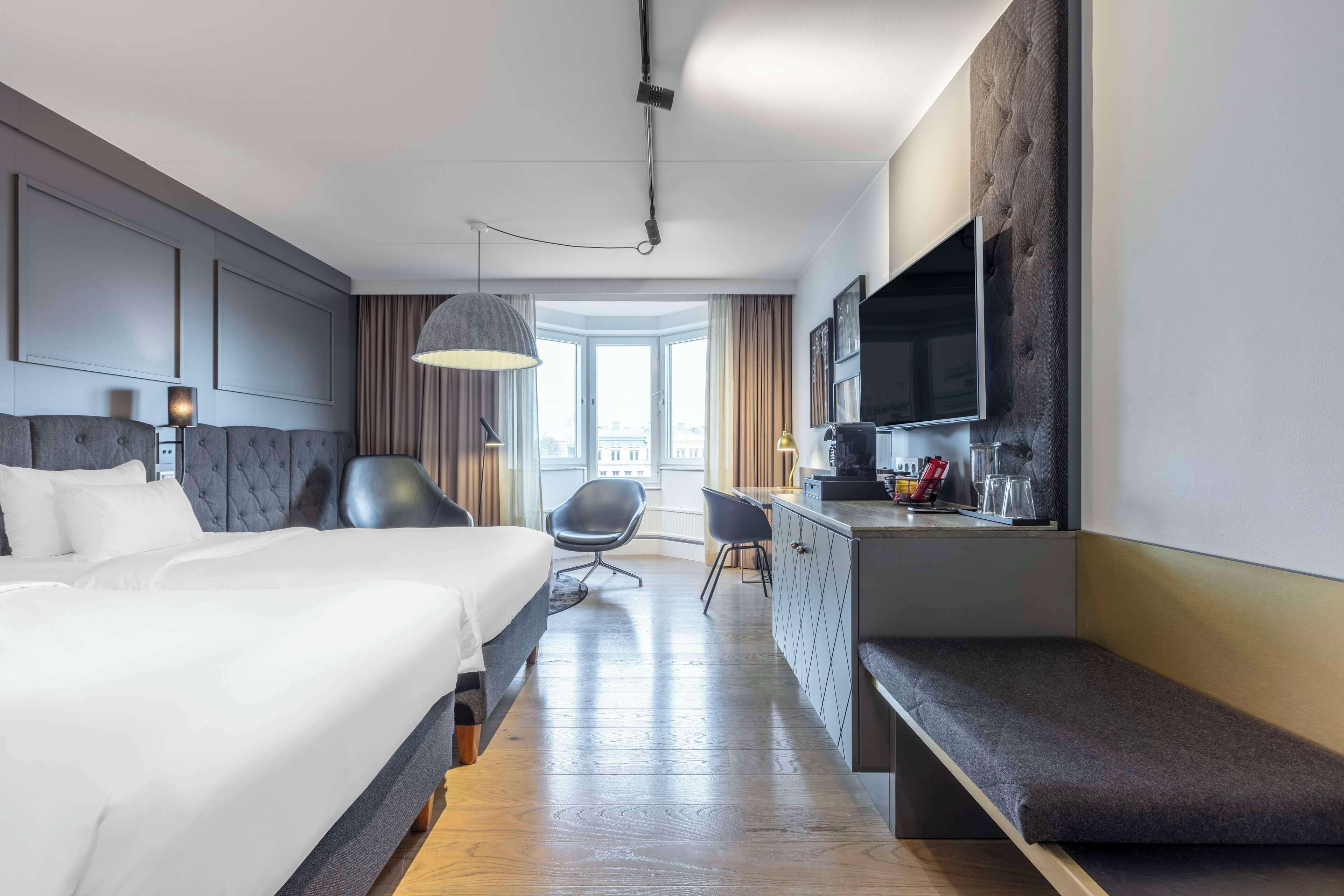 Quarto de hotel moderno com duas camas, uma escrivaninha com televisão de tela plana, um banco e vista para a cidade.
