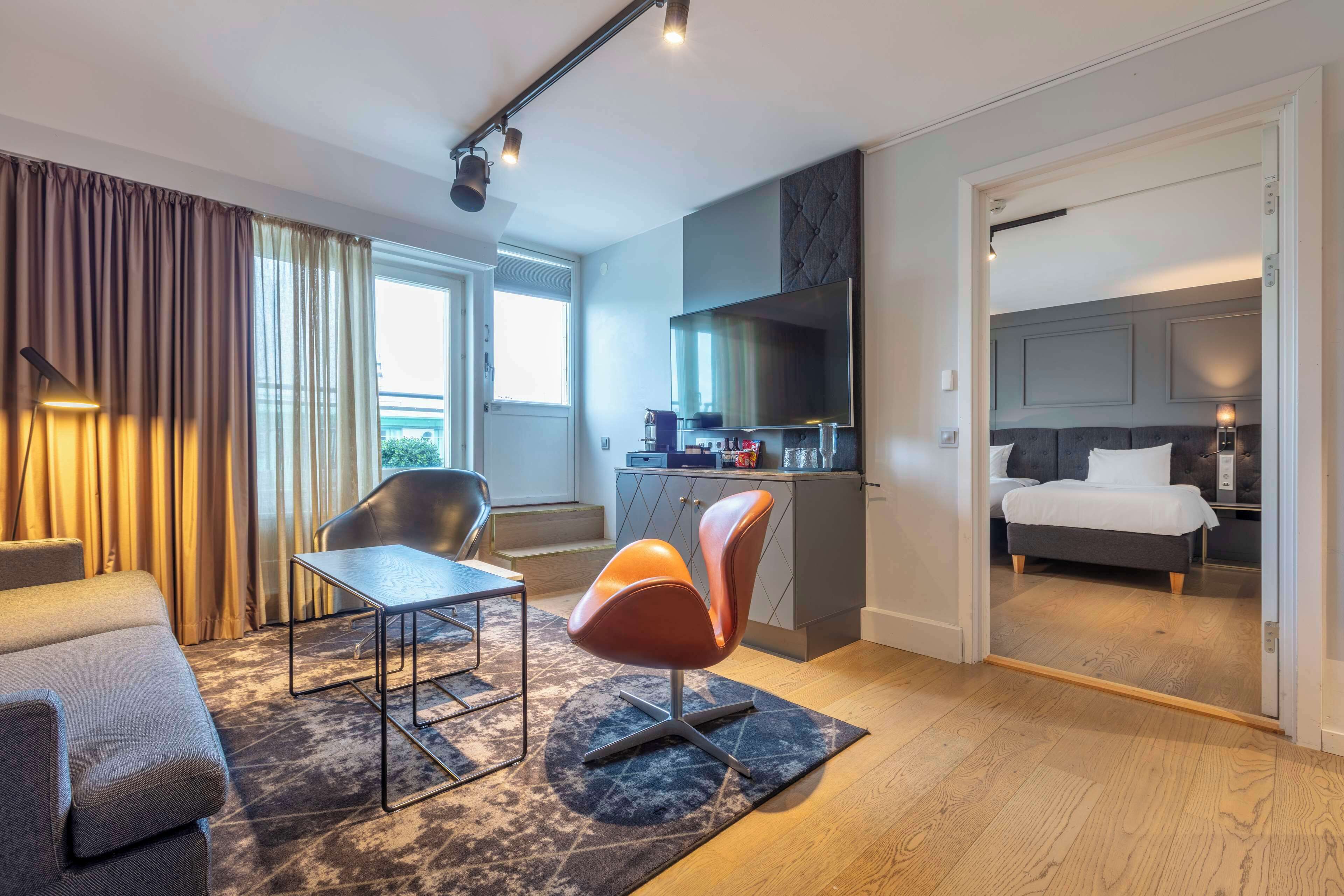 Una habitación de hotel moderna con sofá, mesa de comedor, una silla, televisor, kitchenette y un dormitorio con cama y mesita de noche.