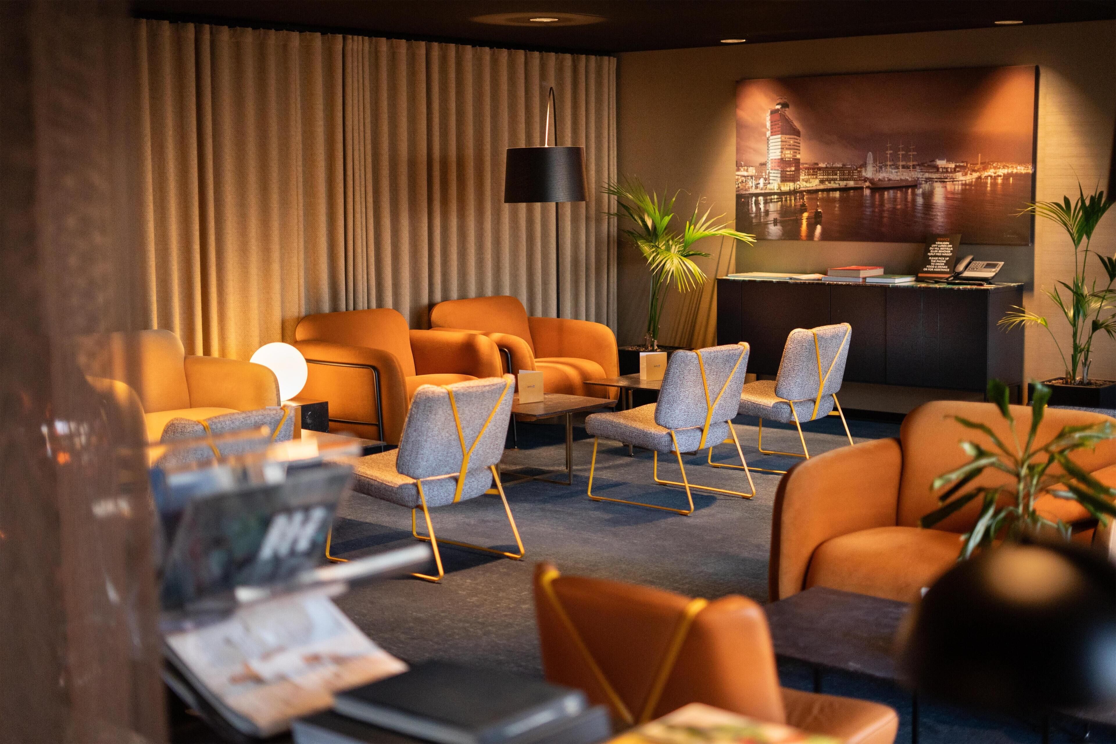 Un hall de hotel con asientos naranja, una recepción, un ventanal con vistas a la ciudad y una planta en maceta.