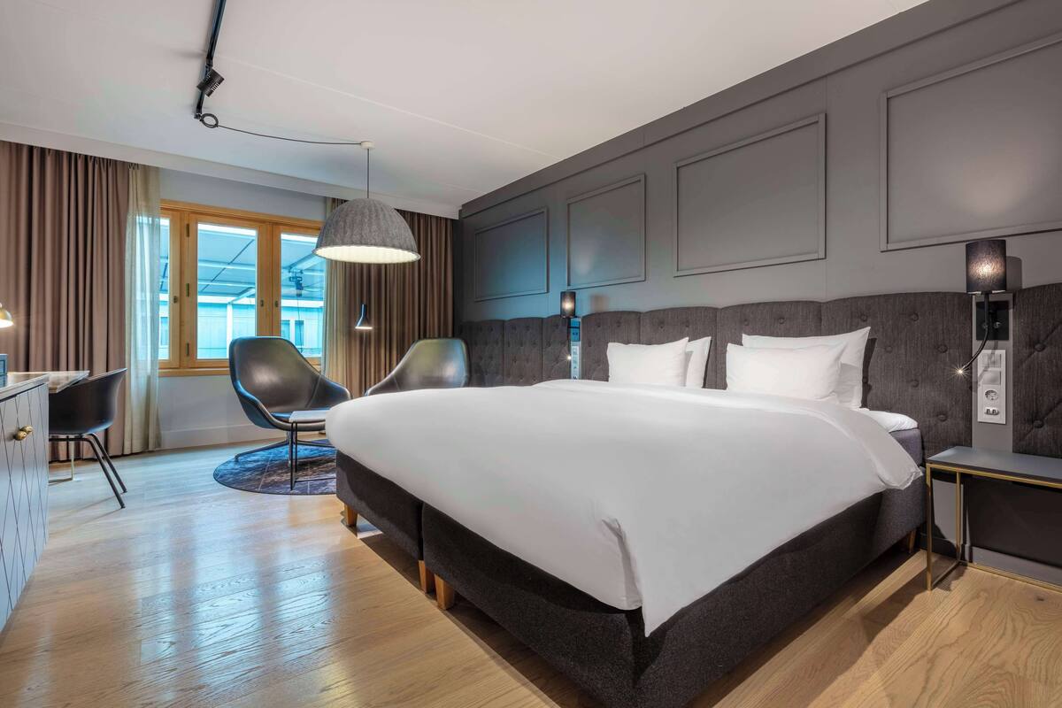A modern hotel room with a large bed, a desk, two chairs, and a view of the outdoors.