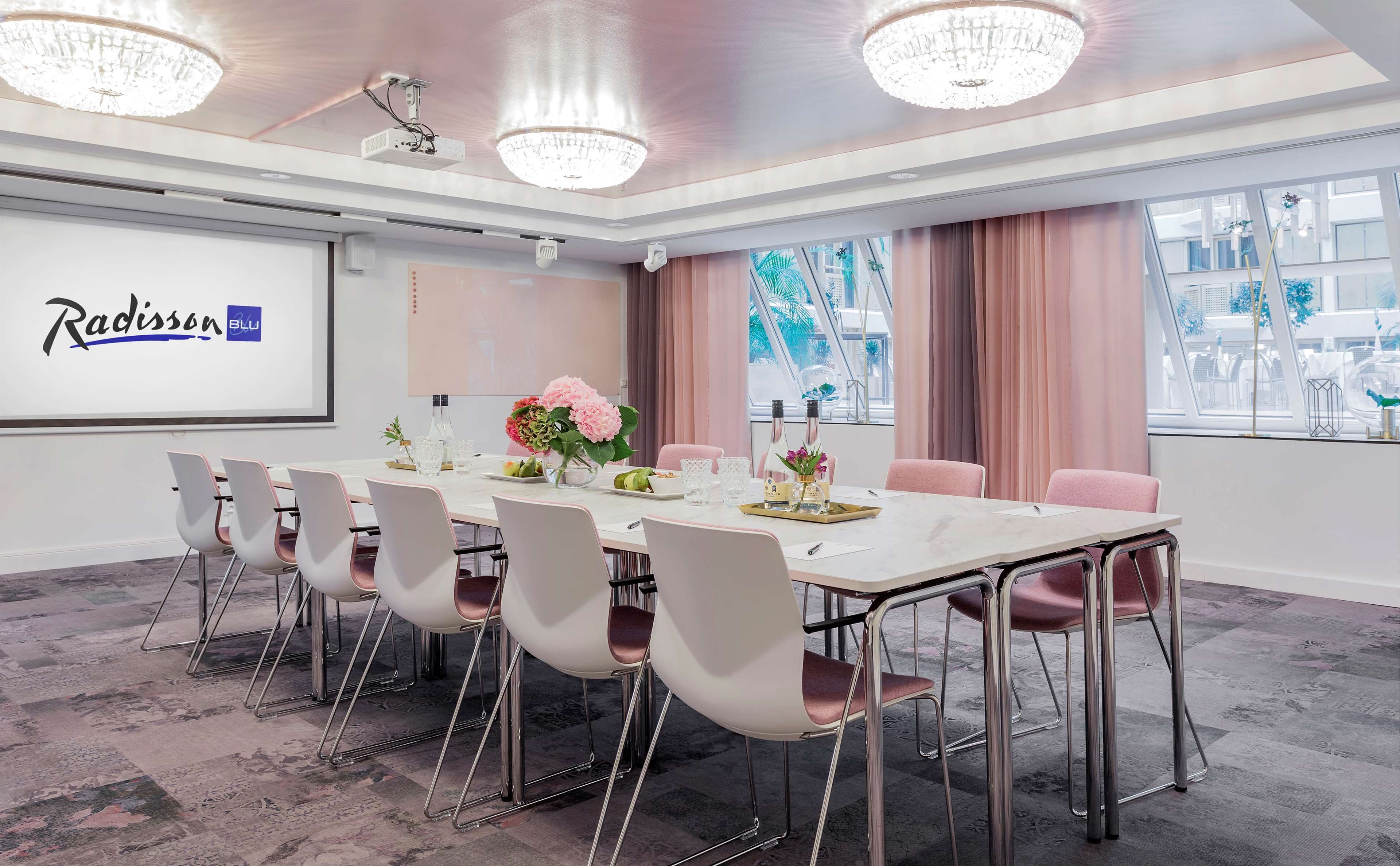 Une salle de conférence avec une table blanche, des chaises roses et un écran de projection affichant le logo « Radisson BLU ».